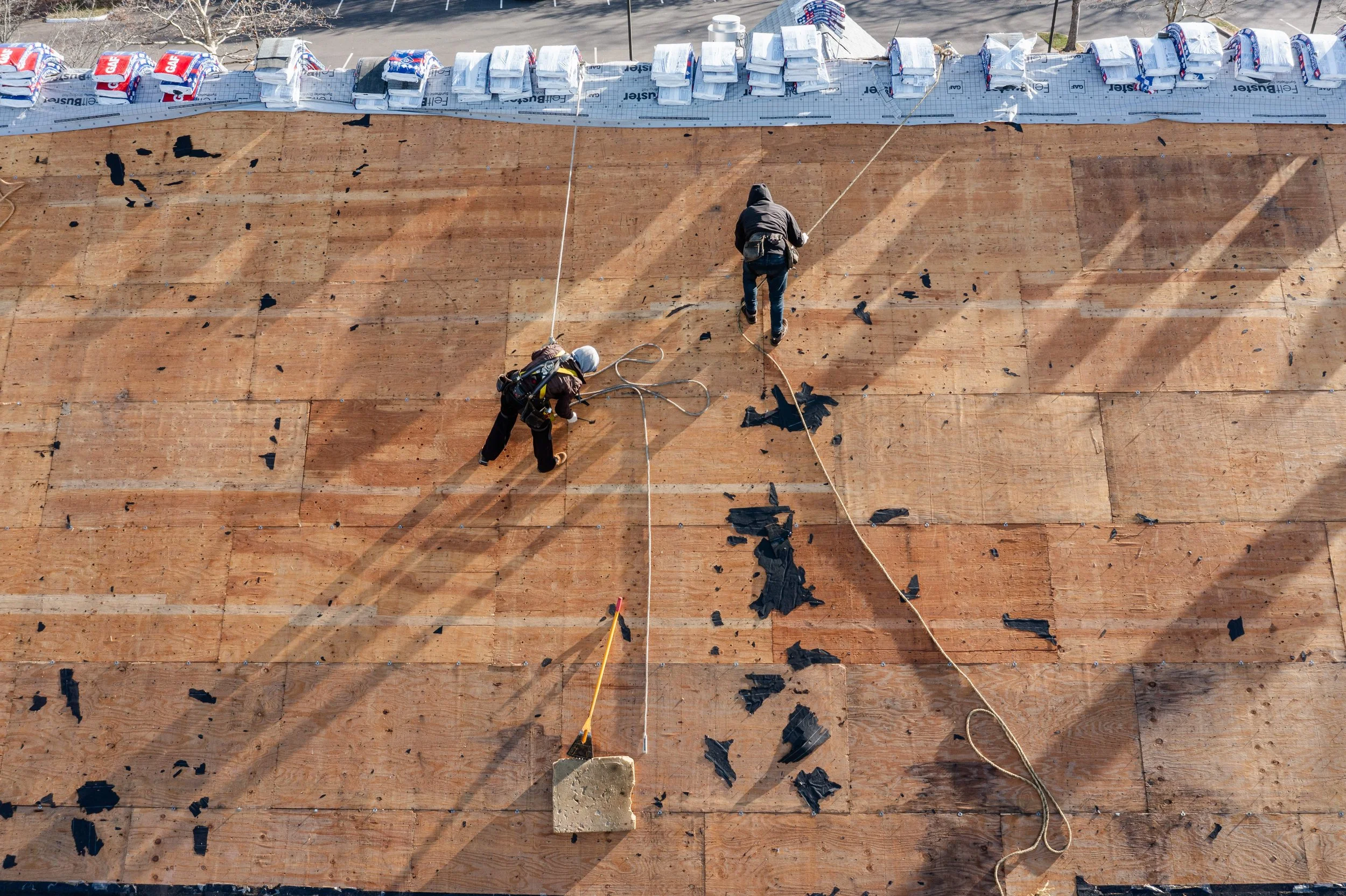 Two construction workers on a wooden roof under construction. One worker is standing while the other is kneeling and appears to be removing old material. There are scattered black debris, tools, and safety equipment around them. The edge of the roof 