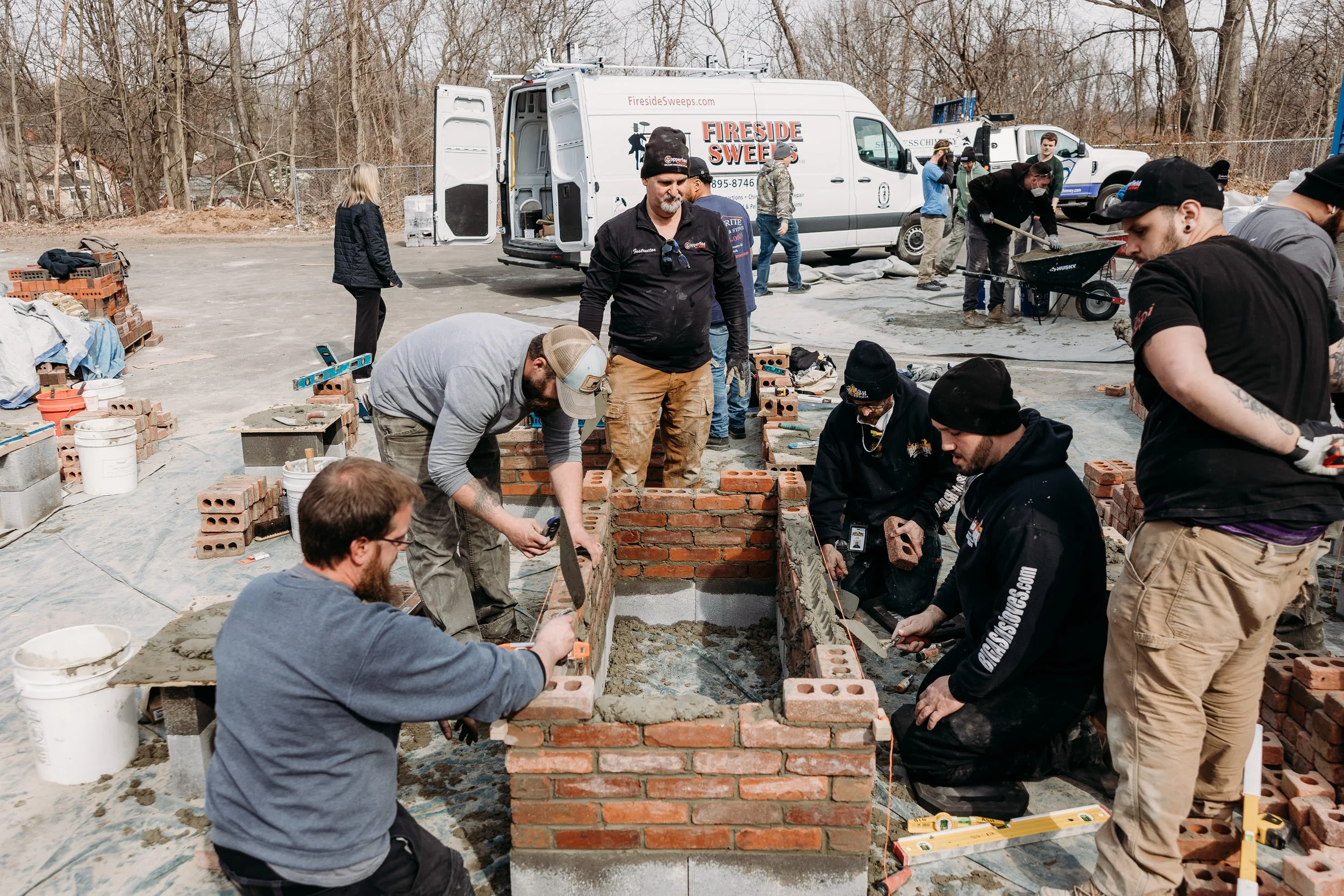 Group of people building a brick structure outdoors, with construction tools and materials, and service vans in the background.