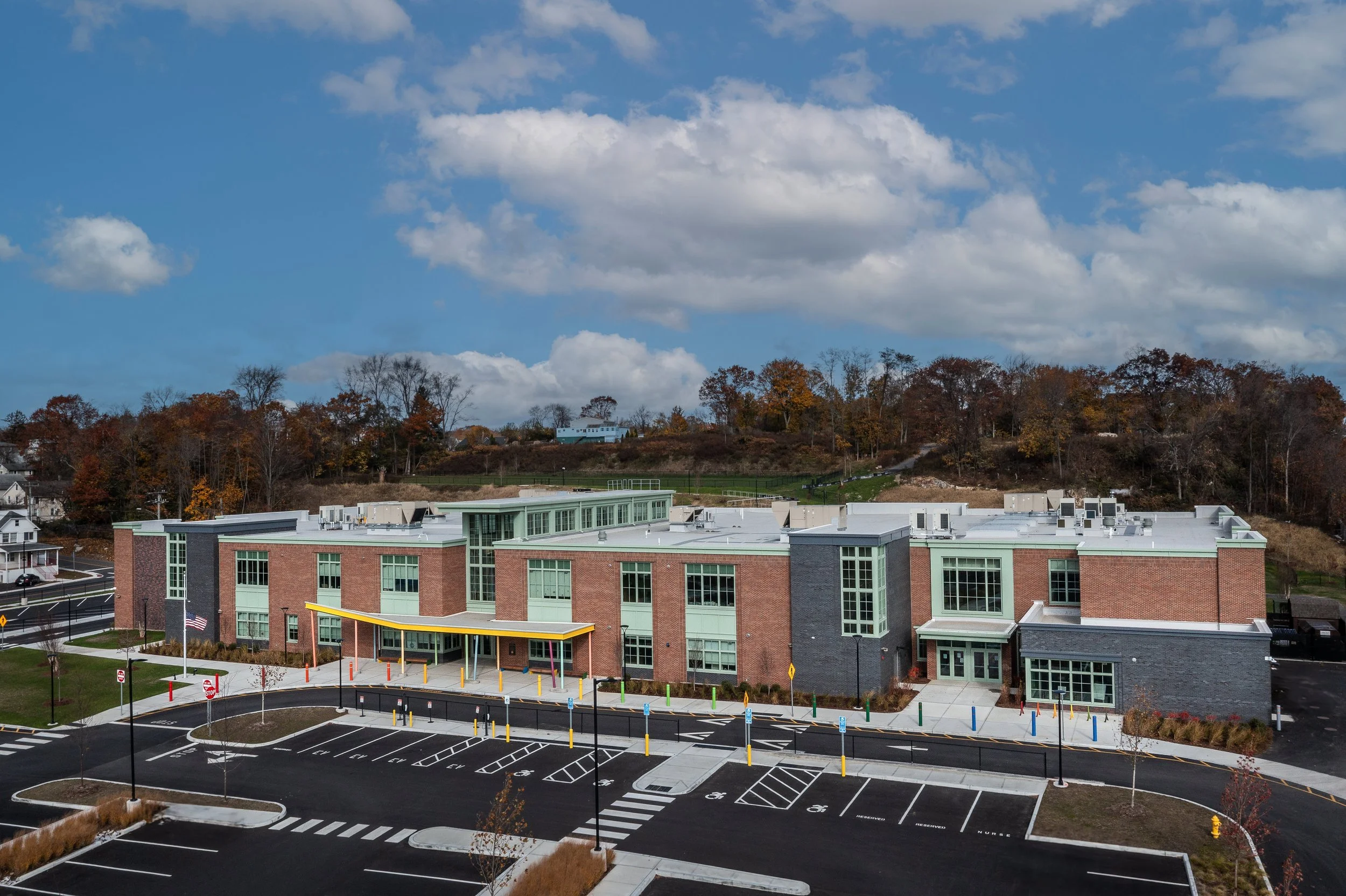An school building with a parking lot in front, surrounded by trees with fall foliage, under a blue sky with clouds.