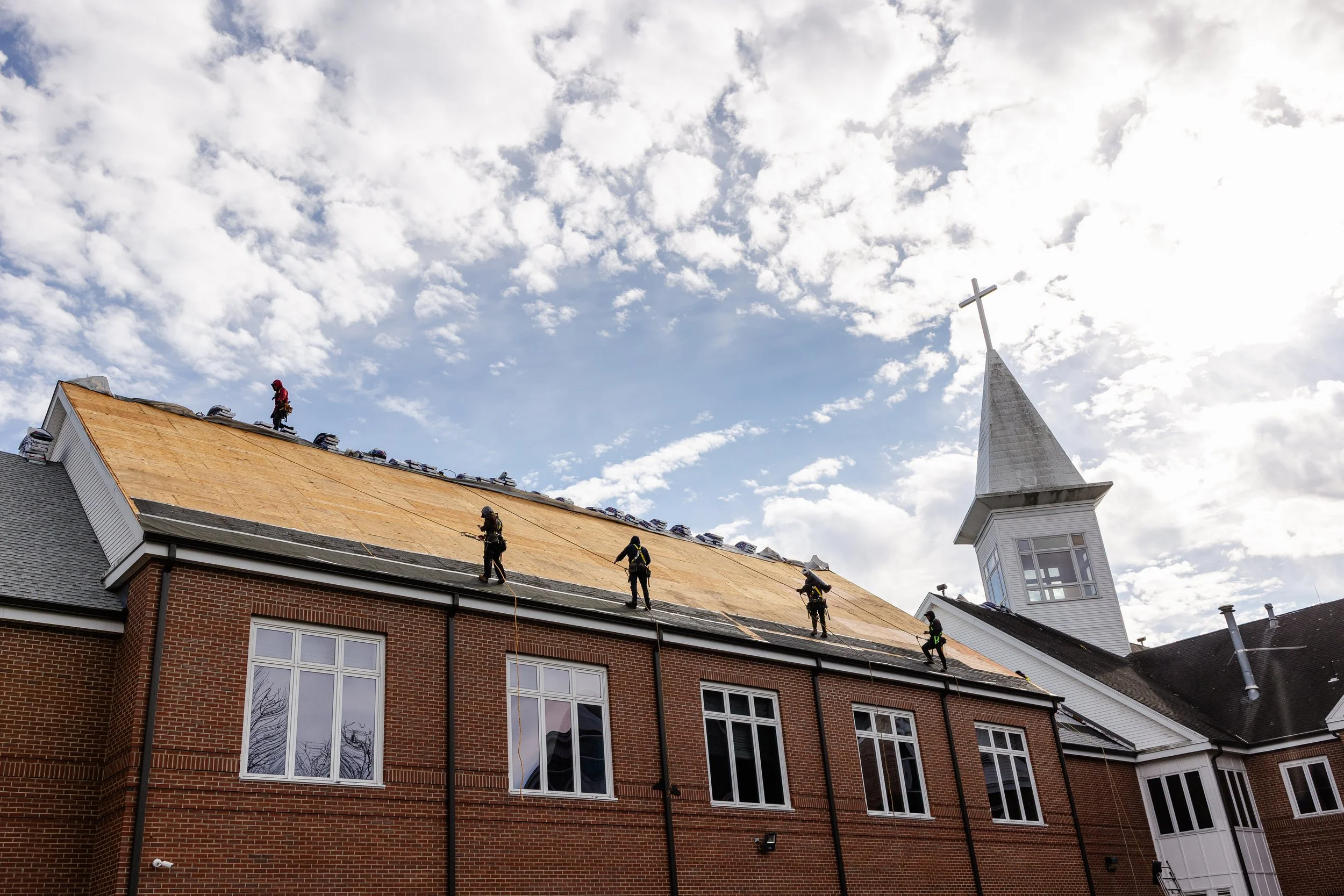 Construction workers installing a new roof on a brick building, with a church steeple and cross in the background, under a partly cloudy sky.