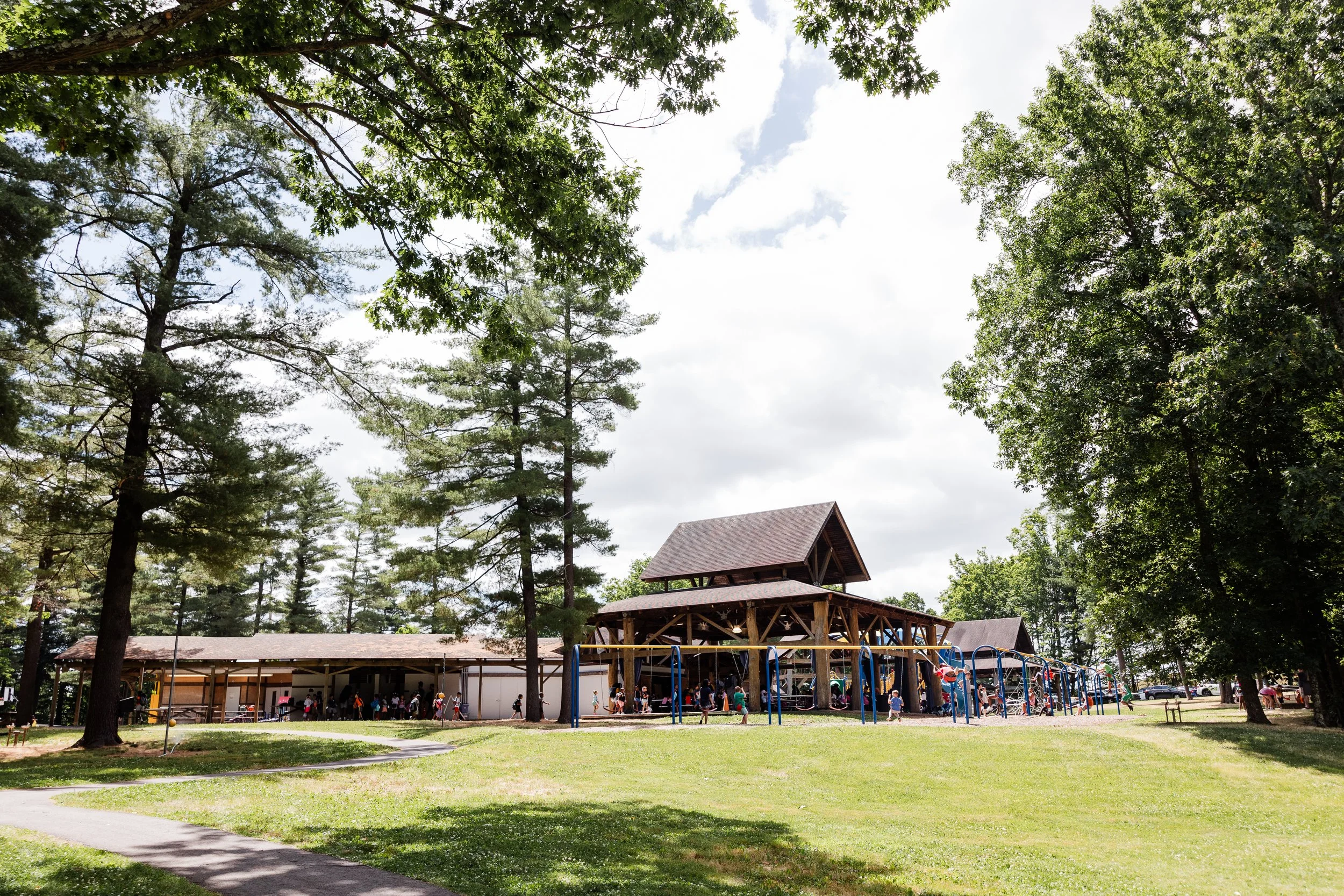 A park with green grass, tall trees, a playground area with swings, and a building in the background with a wooden structure and a peaked roof. Several children and adults are present.