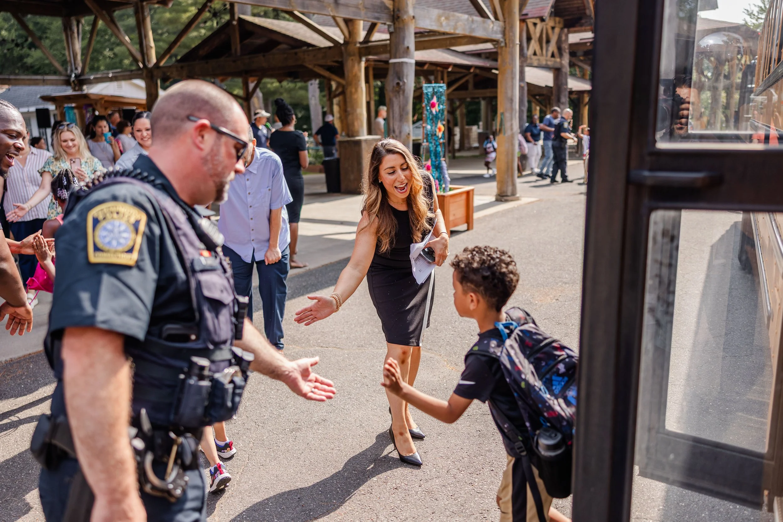 A woman appears to be greeting or saying goodbye to a young boy with a backpack outside a public building, while a police officer stands nearby. Several people are in the background, some smiling and taking photos, in an outdoor environment with wood