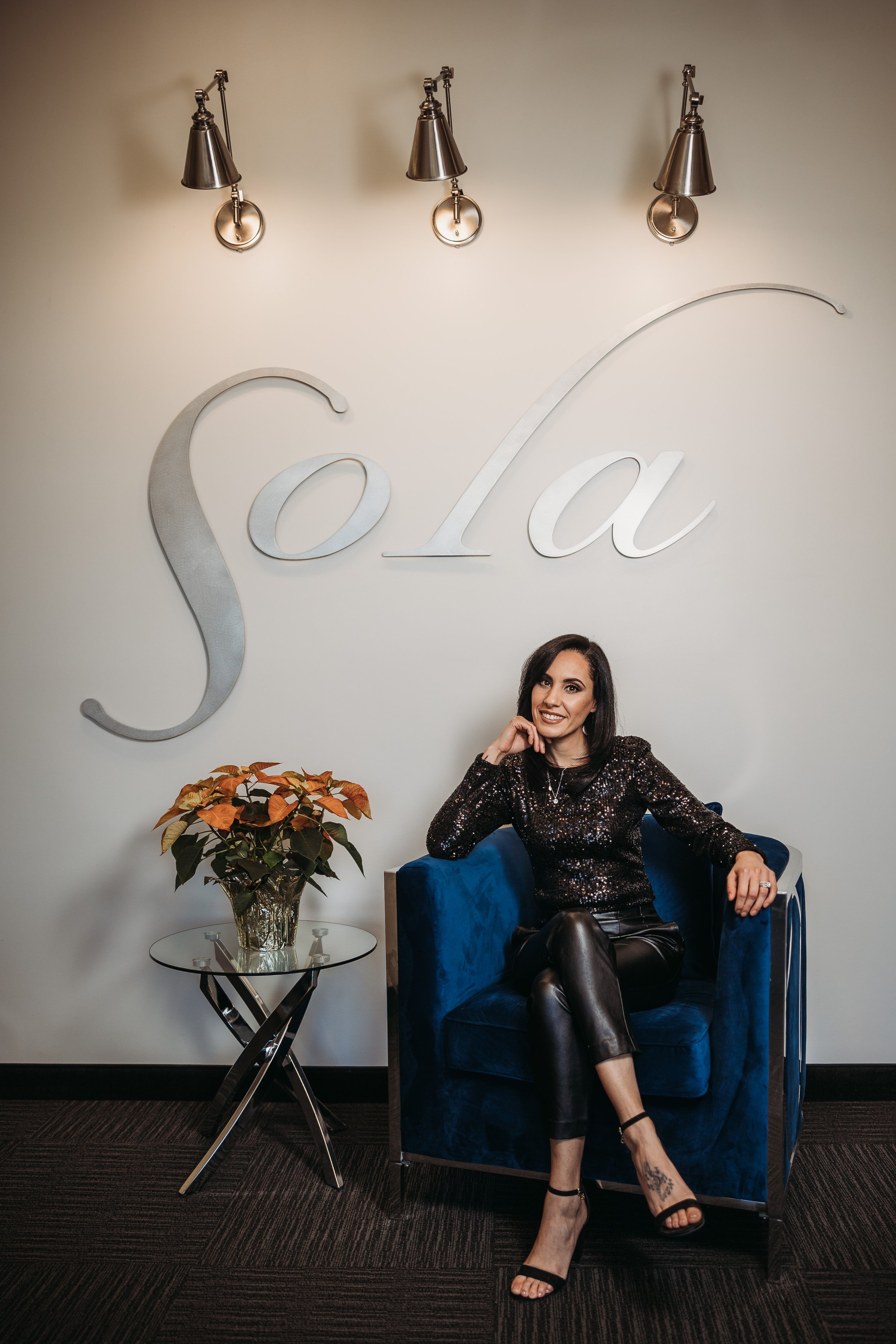A woman sitting in a blue velvet chair with a large silver 'Sola' sign on the wall behind her inside a modern room with three wall lamps and a small glass table with a plant.