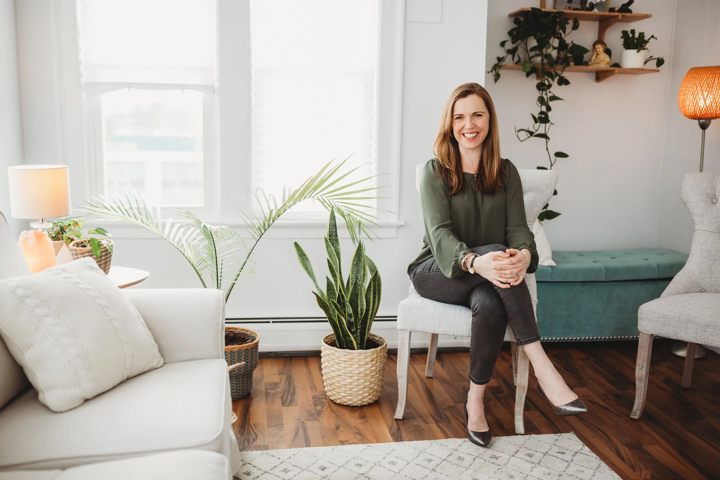 A woman with long brown hair sitting on a white chair in a well-lit living room, smiling at the camera. The room has white walls, large windows, and various indoor plants. There is a white sofa, a green bench, and a wooden shelf with decorative items