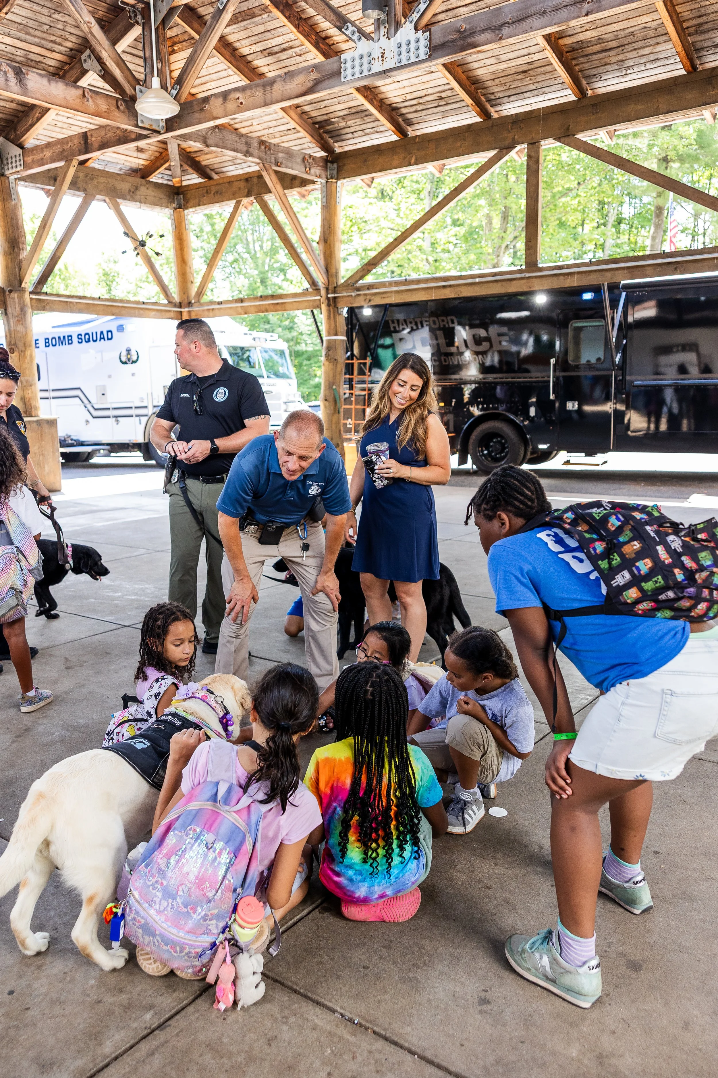 Children and adults gathered indoors with police officers and service dogs, engaging in a community event under a wooden pavilion, with police vehicles in the background.