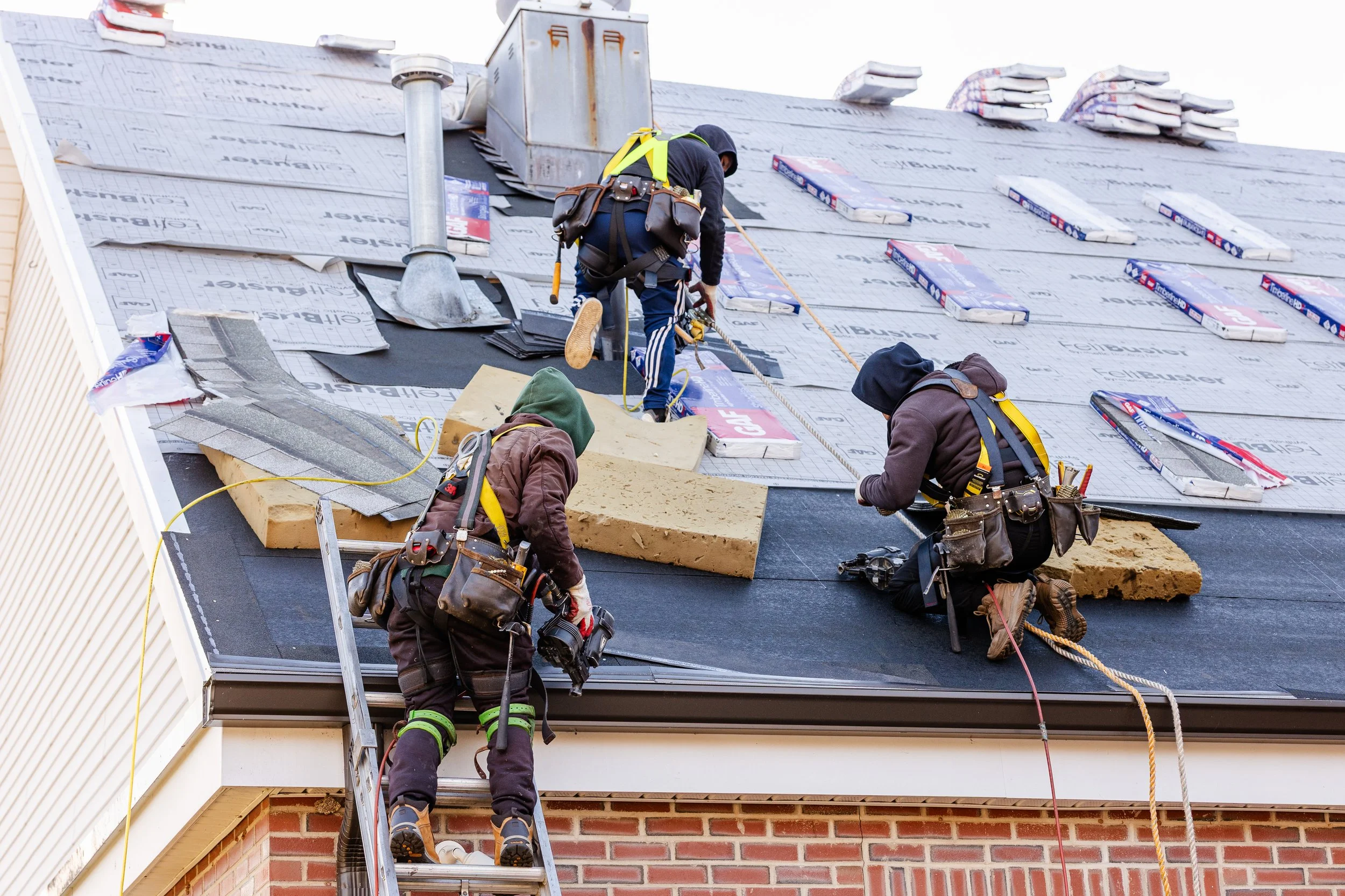 Three construction workers installing or repairing the roofing on a house, working on a steep roof with various materials and tools.