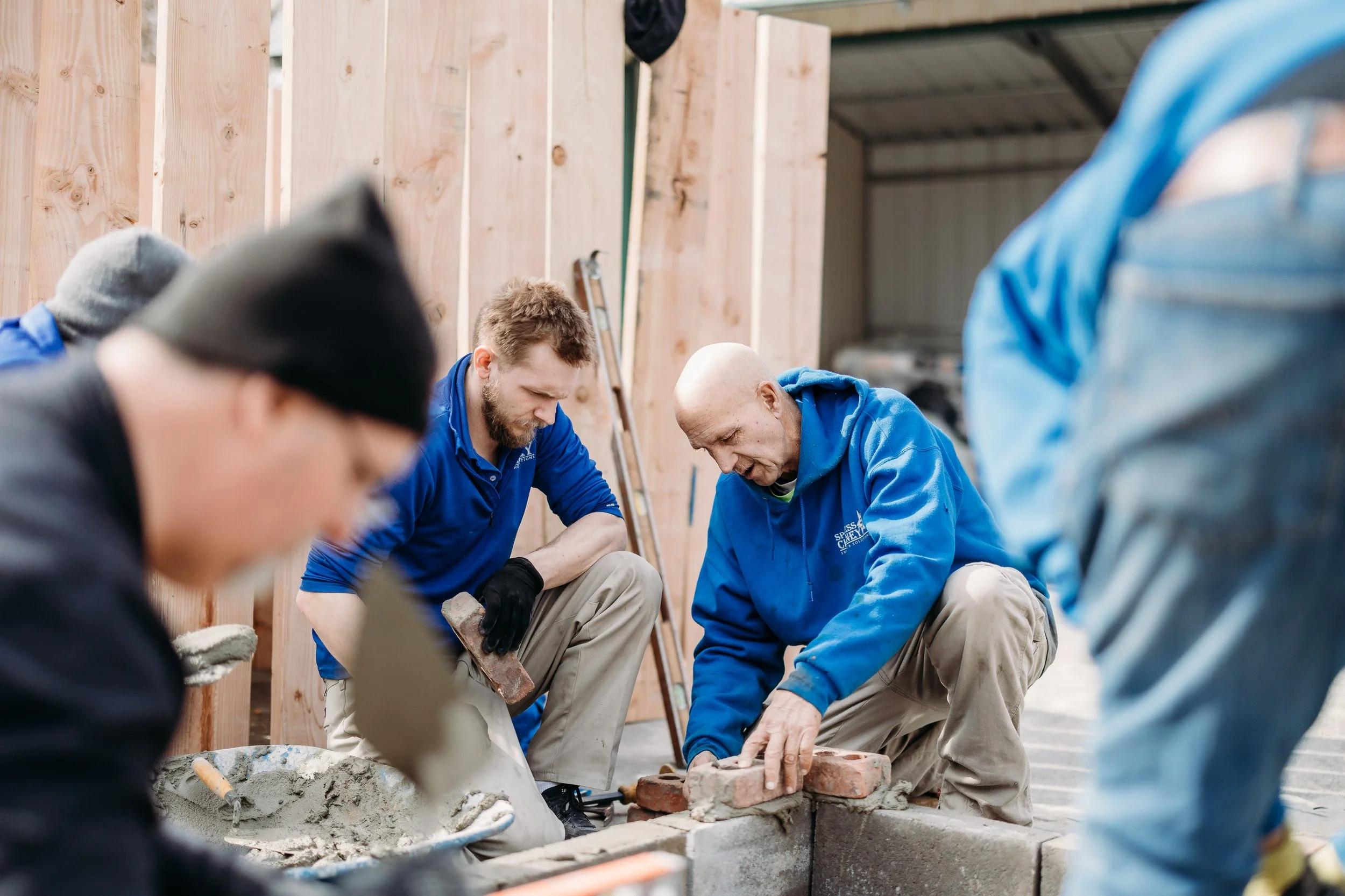Group of men working on construction of a wall, wearing casual clothing and gloves, with tools and building materials on site.