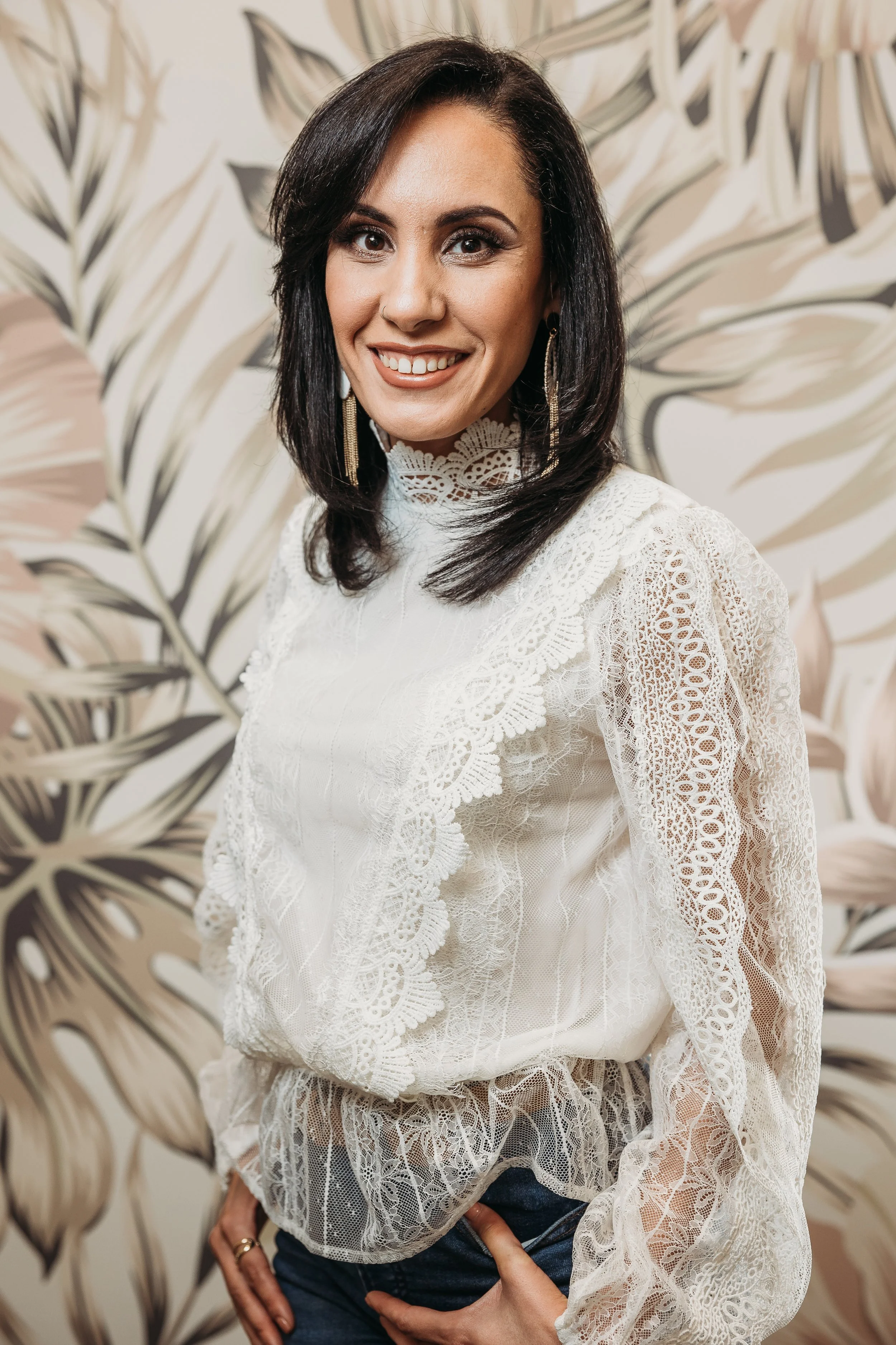 A woman with dark hair styled in loose waves, wearing a white lace blouse with intricate patterns, standing in front of a beige and black botanical wallpaper, smiling at the camera.