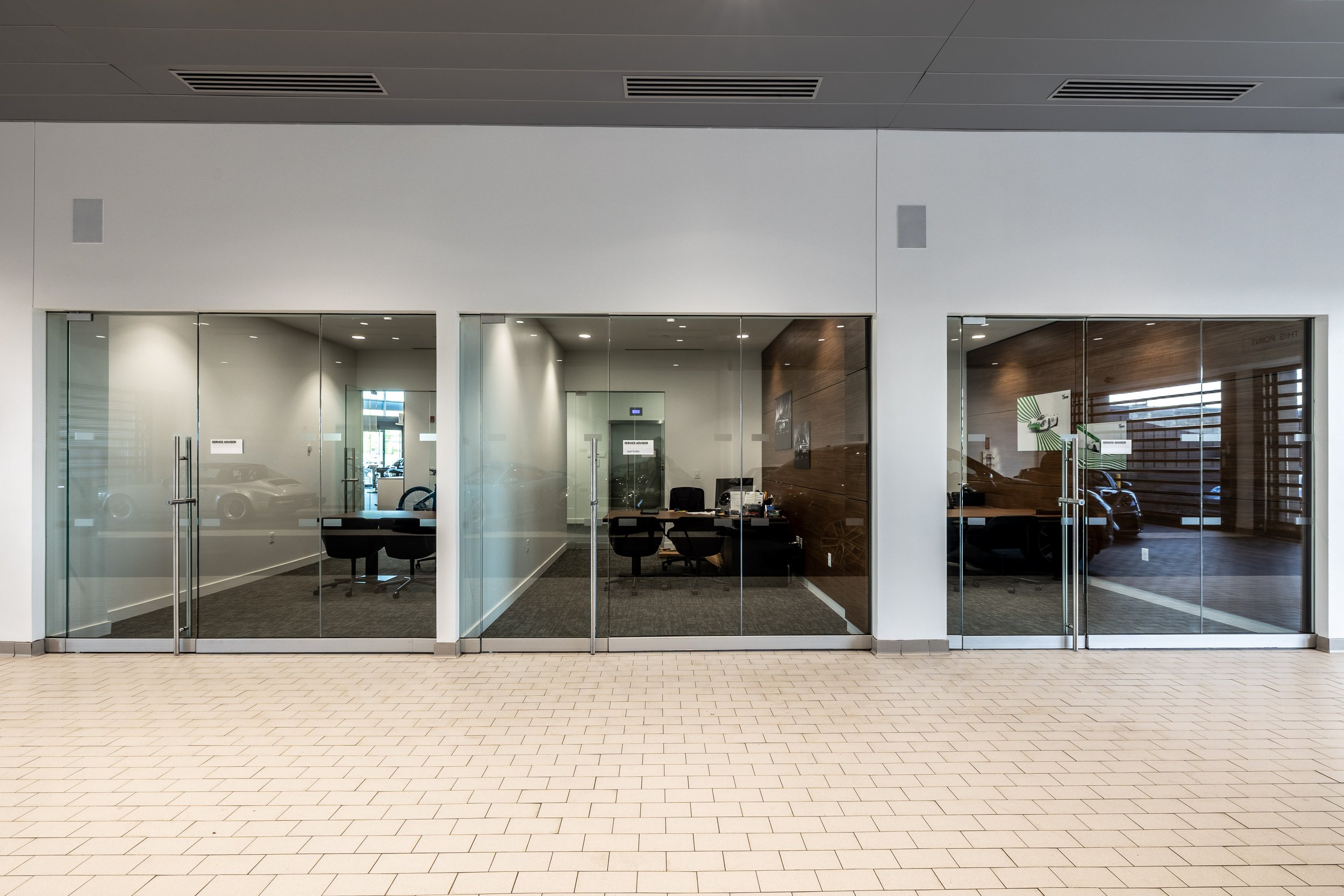 Empty modern office with glass walls, wooden accent wall, black chairs, desk with computers, and parking lot visible through windows.
