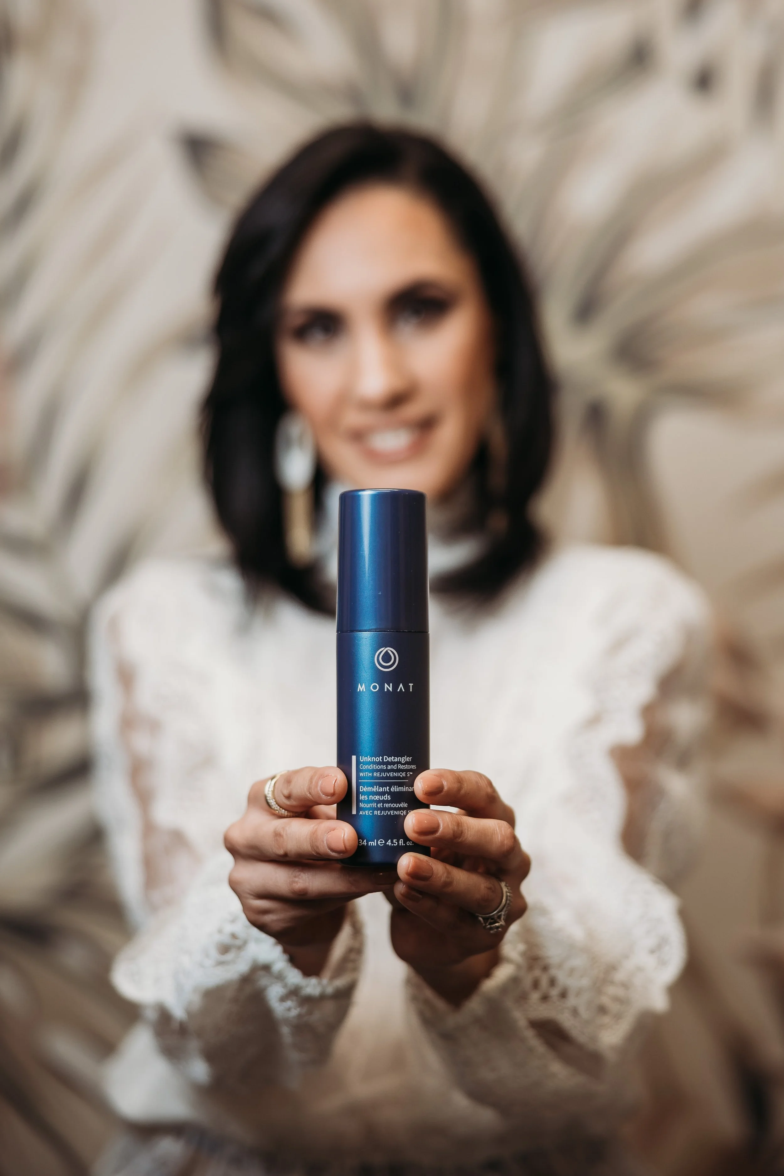 A woman with dark hair holding a blue bottle of Monat skincare product centered in front of her, with a blurred background.