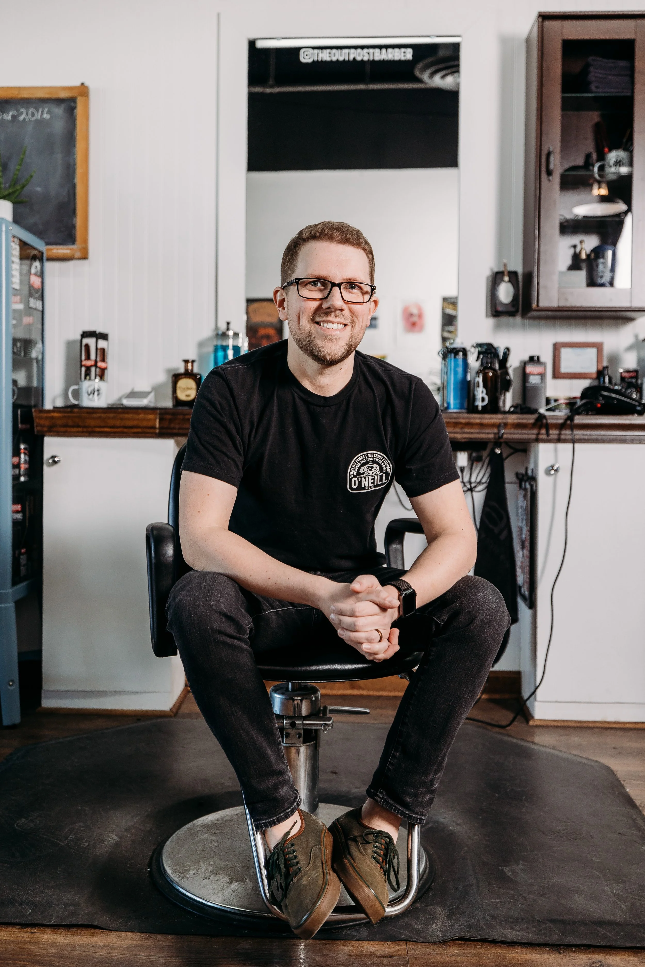 A man with glasses, short hair, and a beard sitting on a barber chair in a barbershop, smiling at the camera.