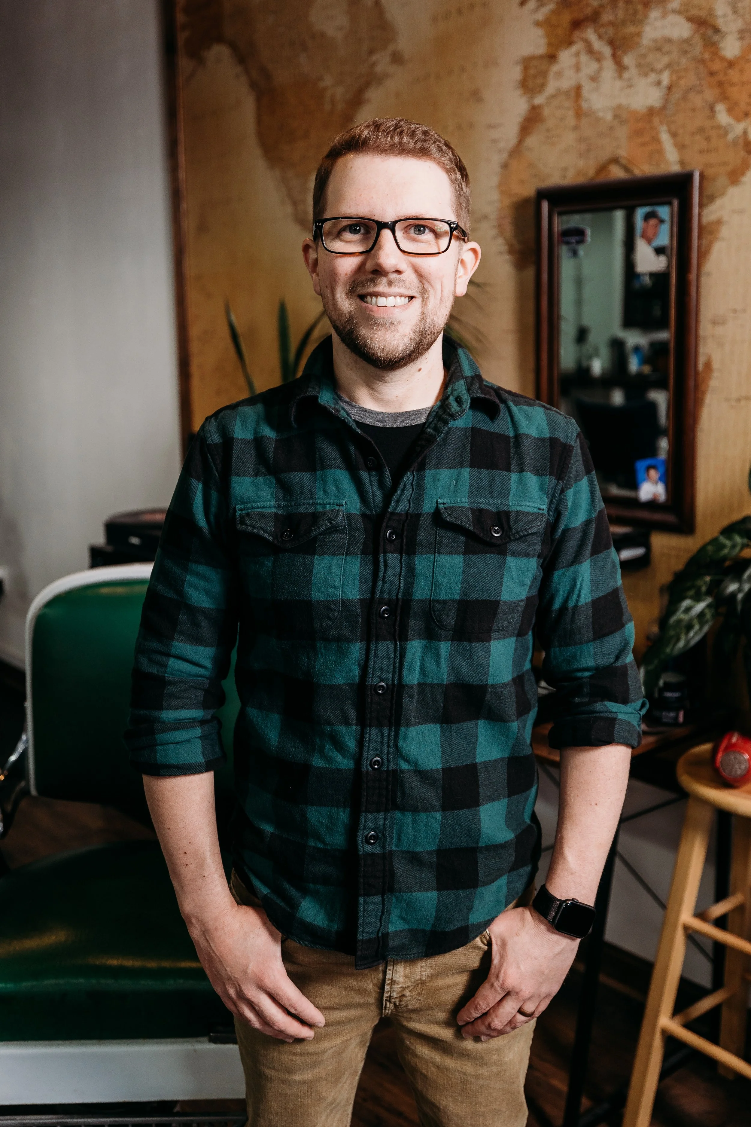 A smiling man with glasses and a beard, wearing a green and black plaid shirt, standing indoors in front of a wall with a world map and a mirror.