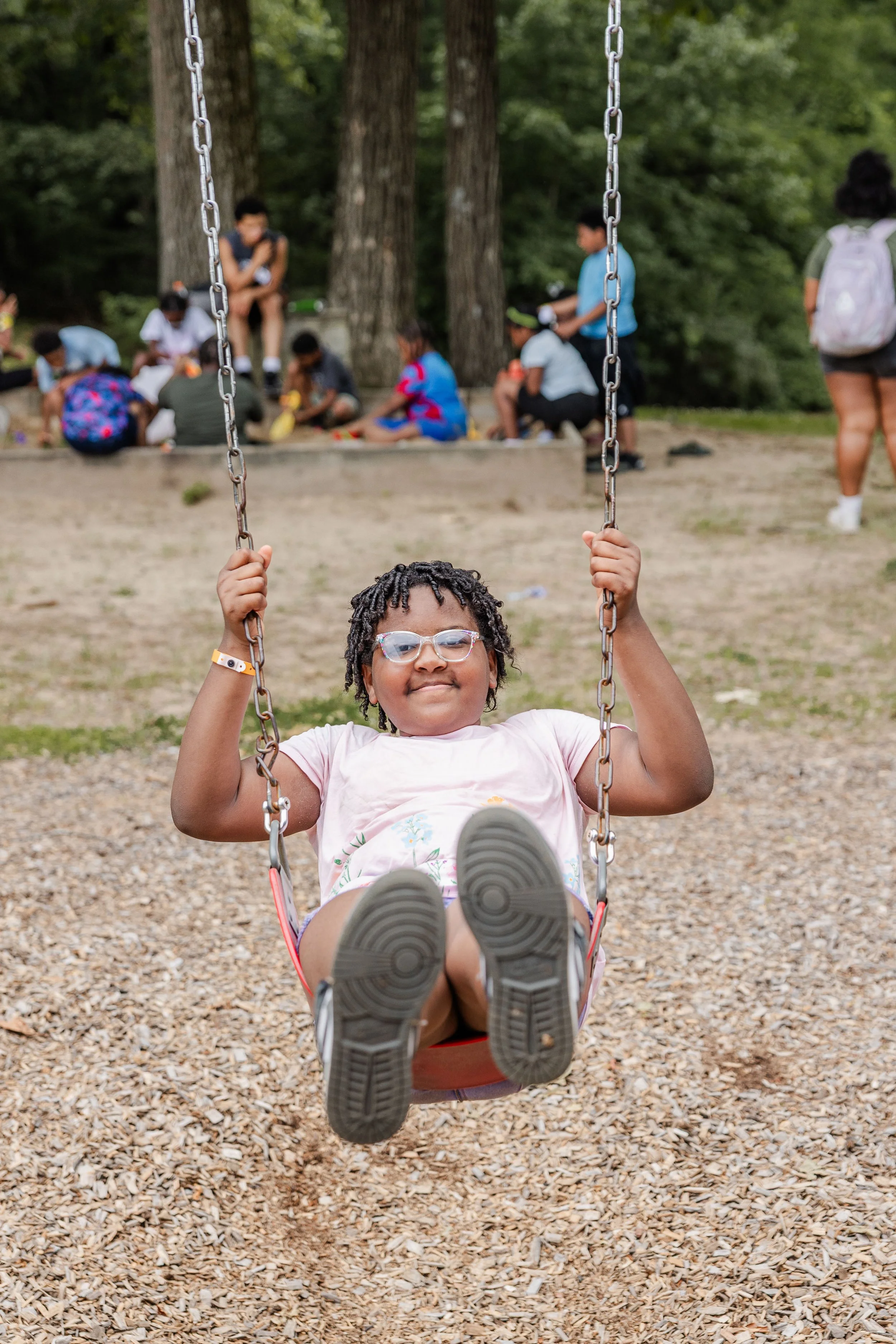 A young girl in glasses and a pink shirt swinging on a playground swing set with a forest in the background and a group of children and adults sitting and standing nearby.