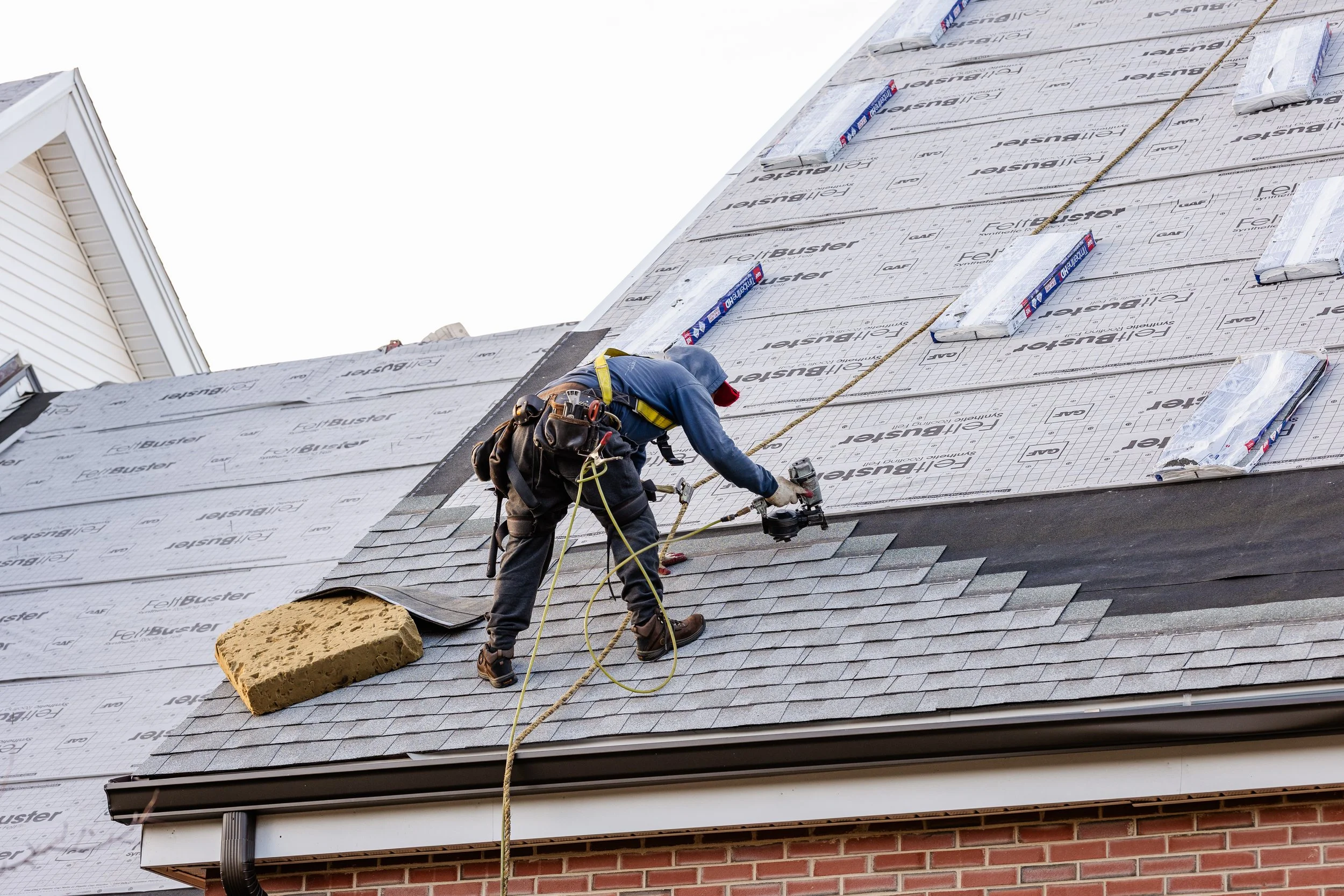 A worker on a roof installing shingles with a nail gun, wearing safety gear, with roofing materials and a brick wall below.