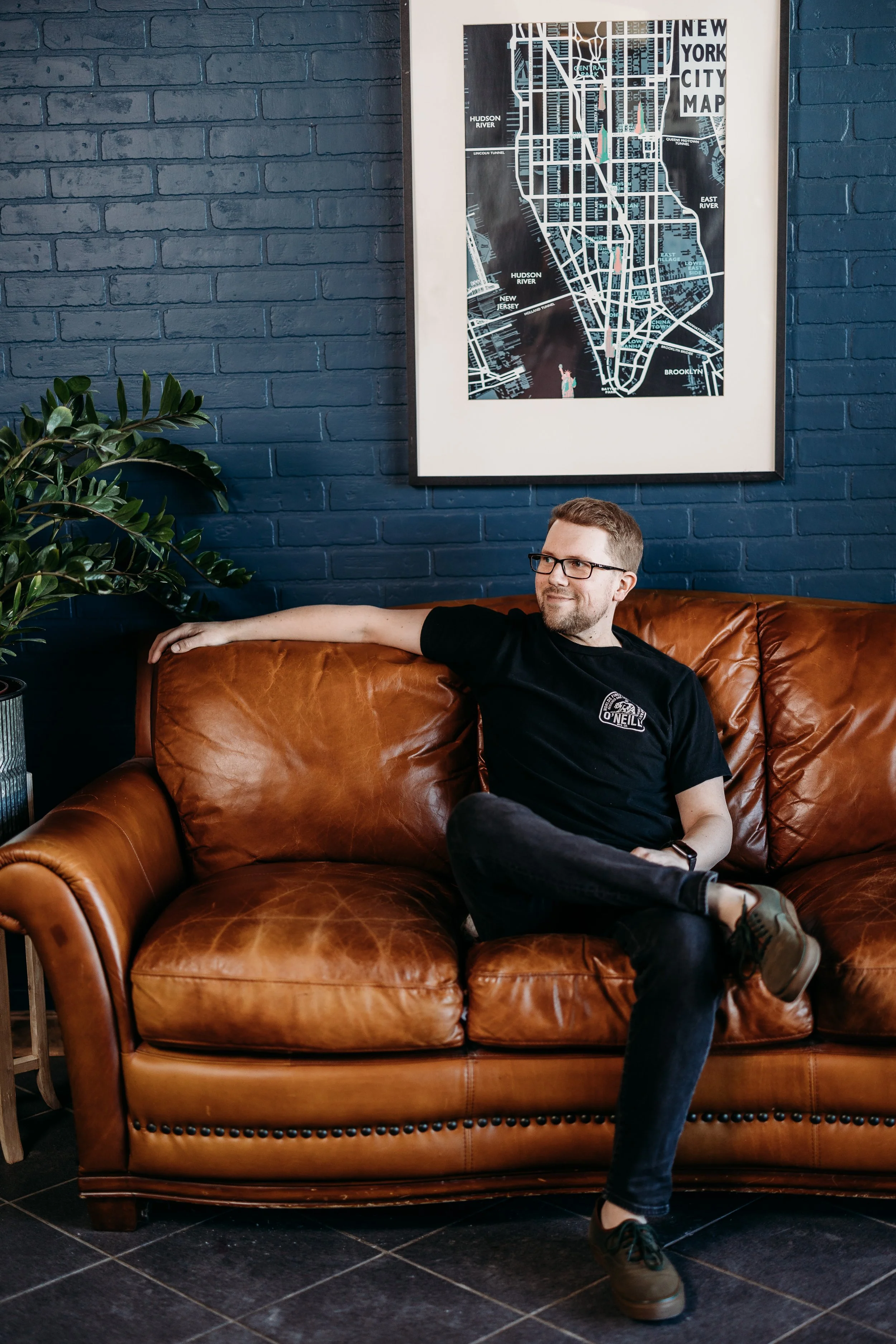 A man with glasses sits on a brown leather couch with a relaxed pose, one arm stretched out on the back of the couch, in a room with a dark blue brick wall, a potted plant, and a framed map of New York City.