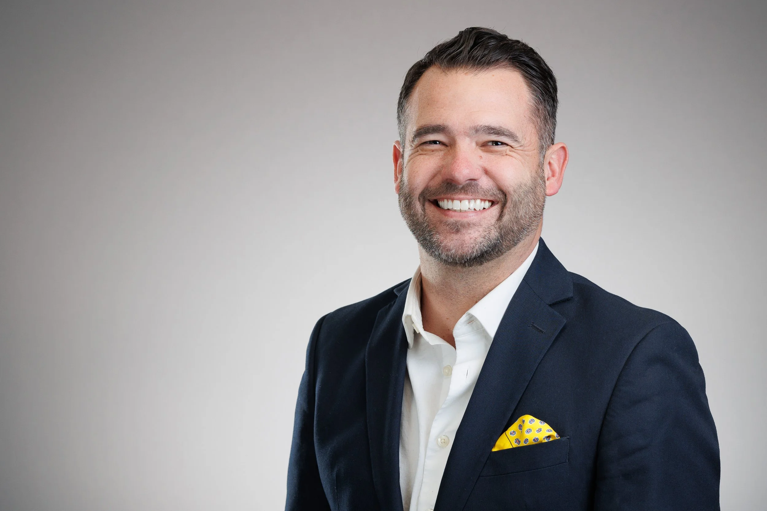 Headshot of a smiling man in a navy blazer and white shirt, with a yellow pocket square, against a plain gray background.