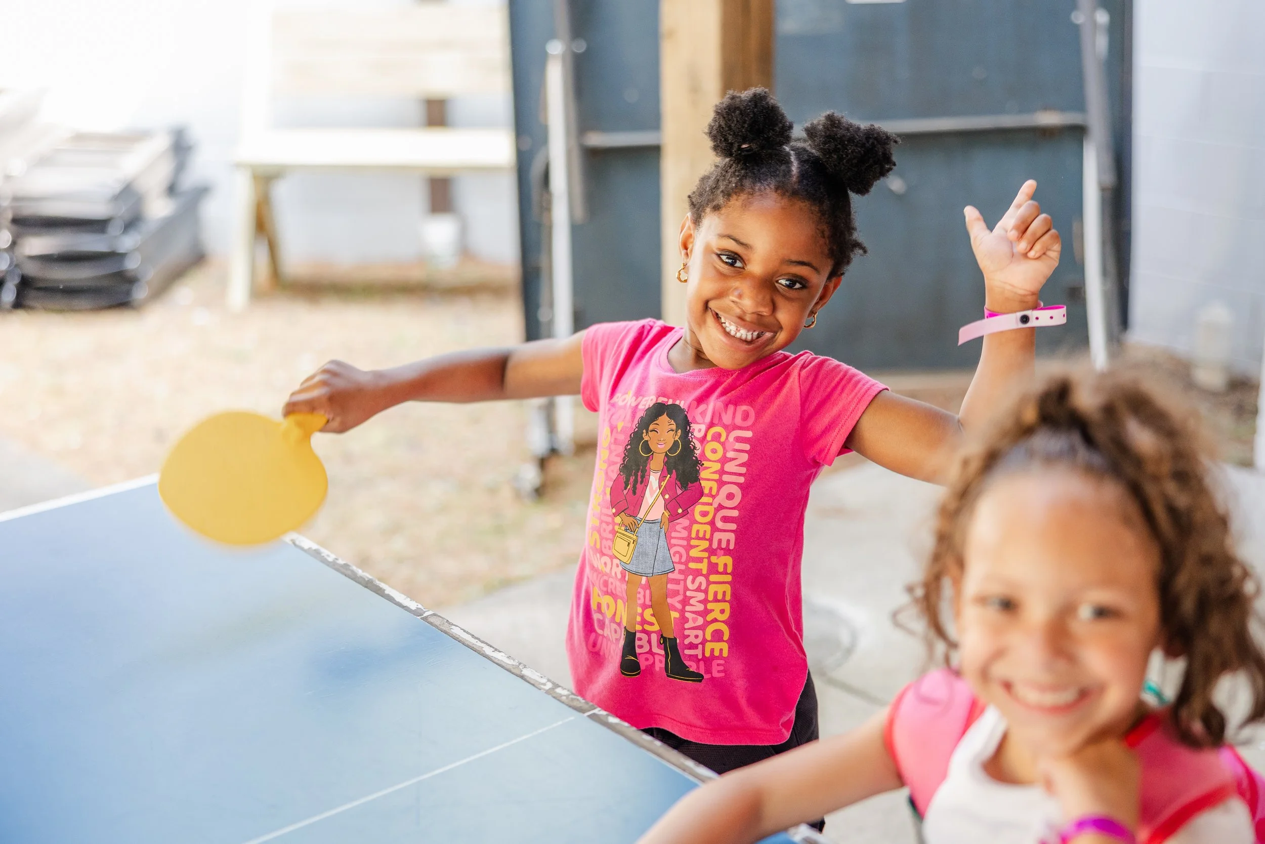 Two young girls playing table tennis outdoors, one with a yellow paddle and the other smiling and wearing a pink shirt.