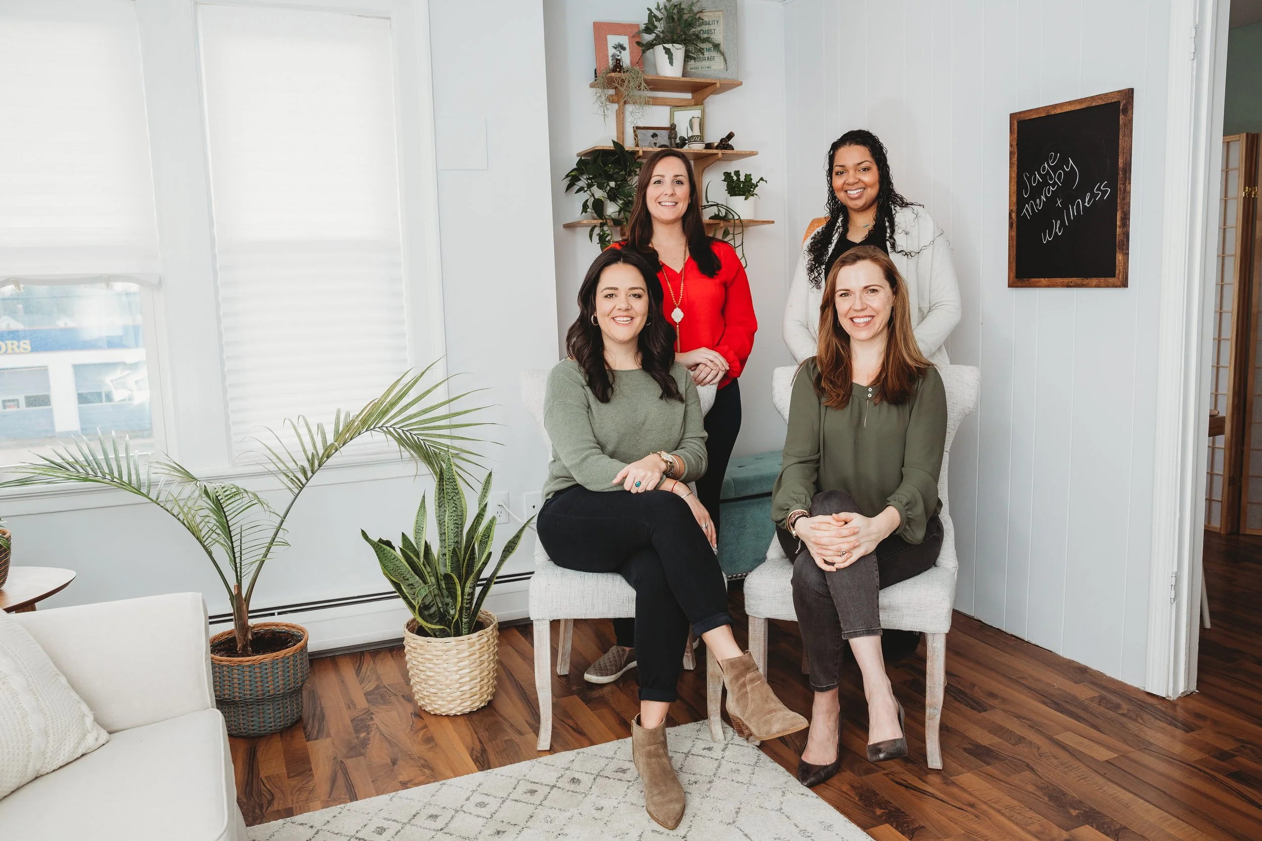 Five women smiling in a bright, modern room with plants, shelves, and a chalkboard reading "Soap, Menopause, Wellness" in the background.