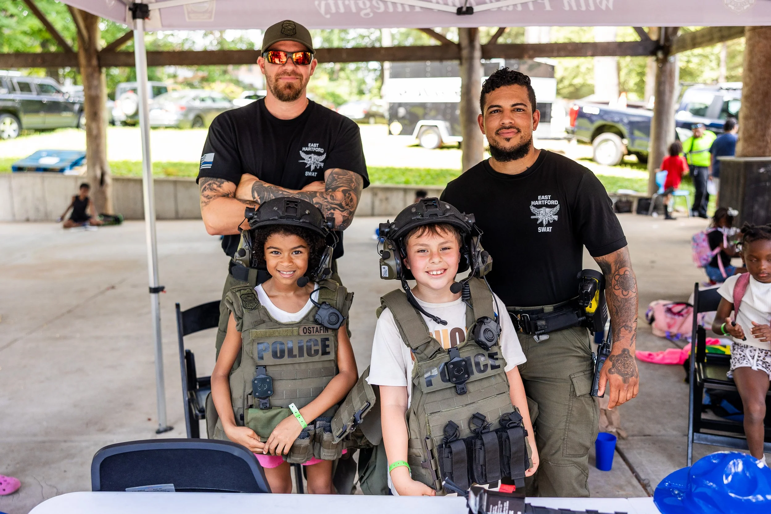 Two police officers and two young girls wearing police vests and helmets, posing under a canopy at an outdoor community event.