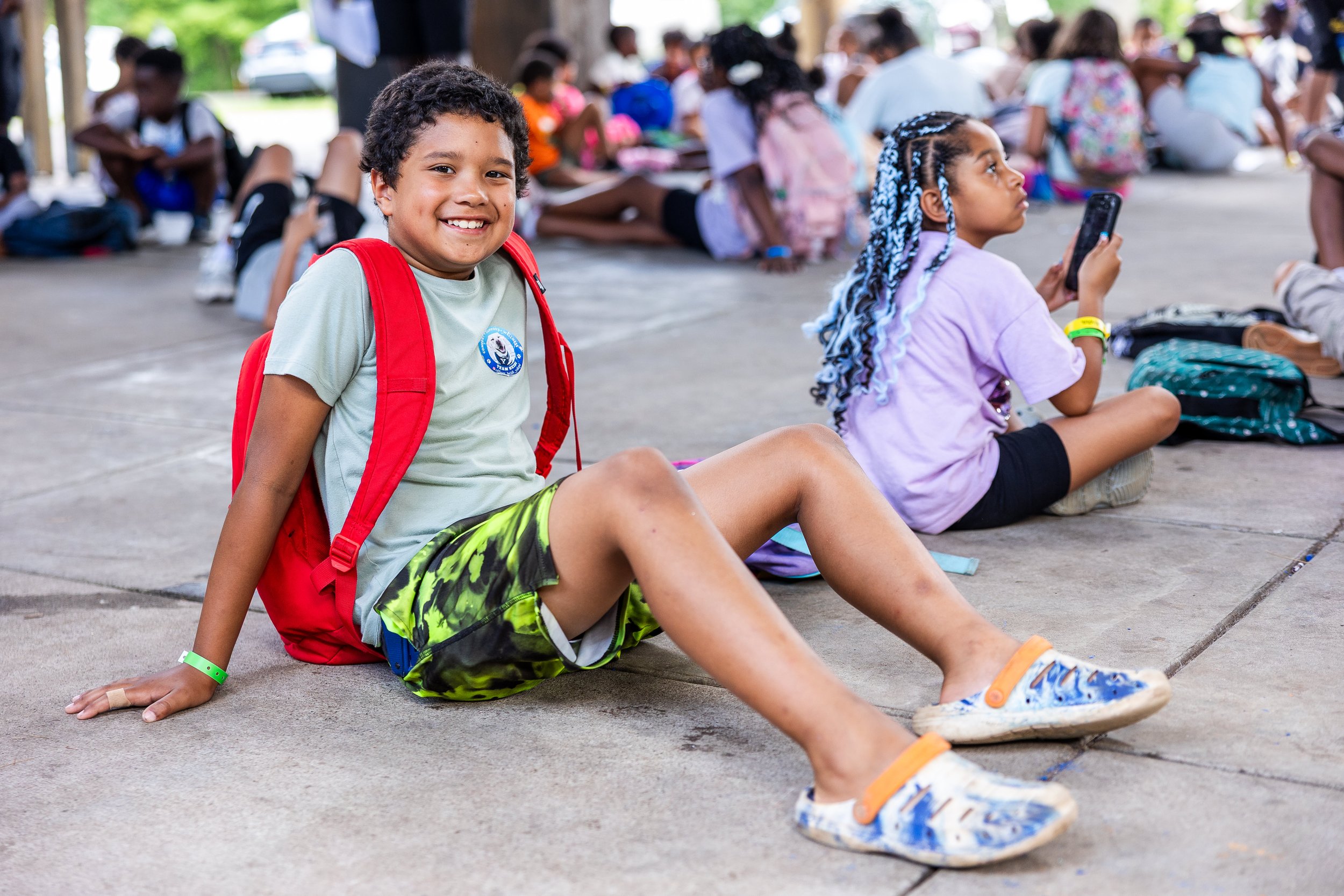A boy sitting on the ground with a red backpack, smiling at the camera, surrounded by other children sitting on the ground indoors or in a covered outdoor area.