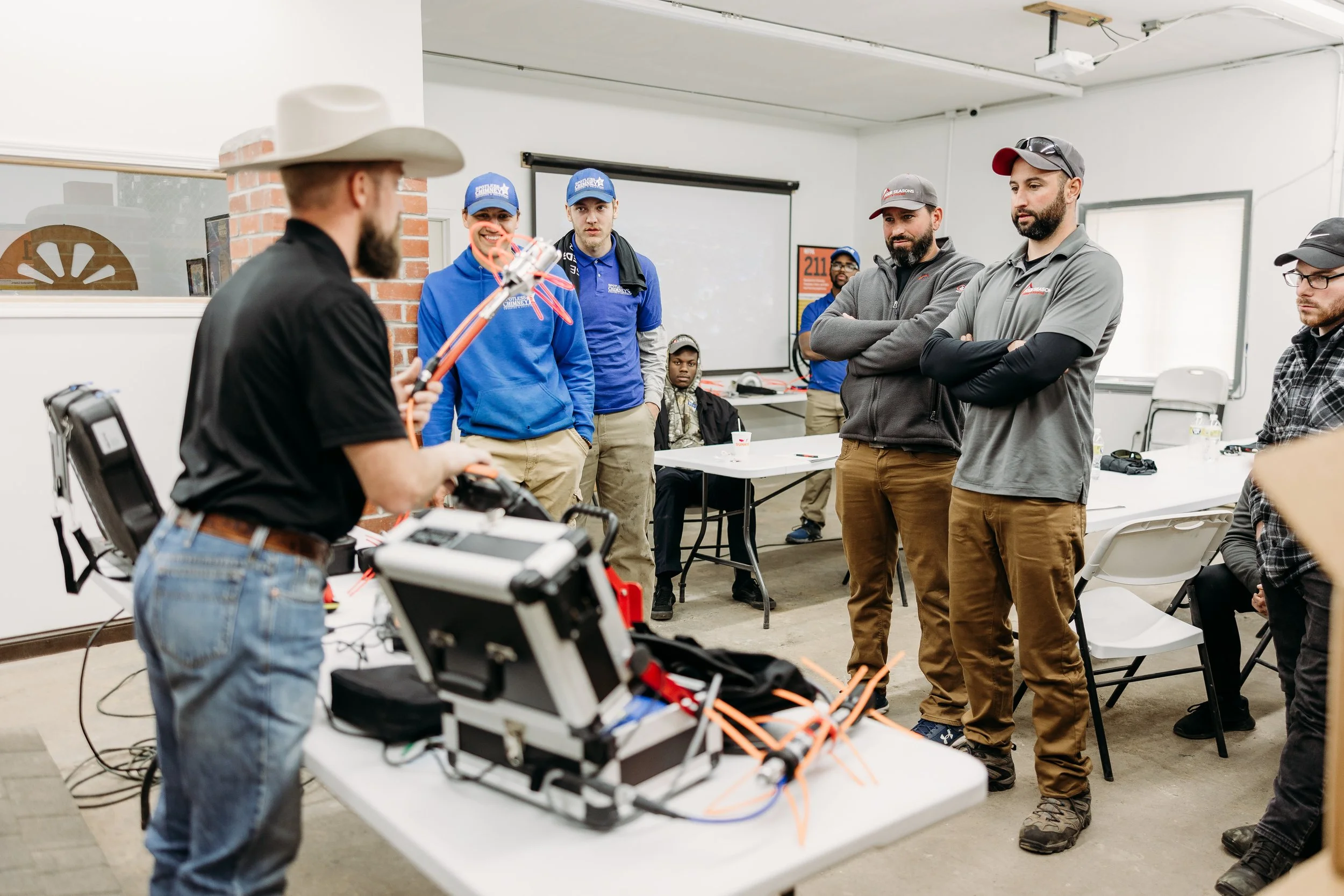 A man in a cowboy hat demonstrates drone technology to a group of people in a classroom setting.