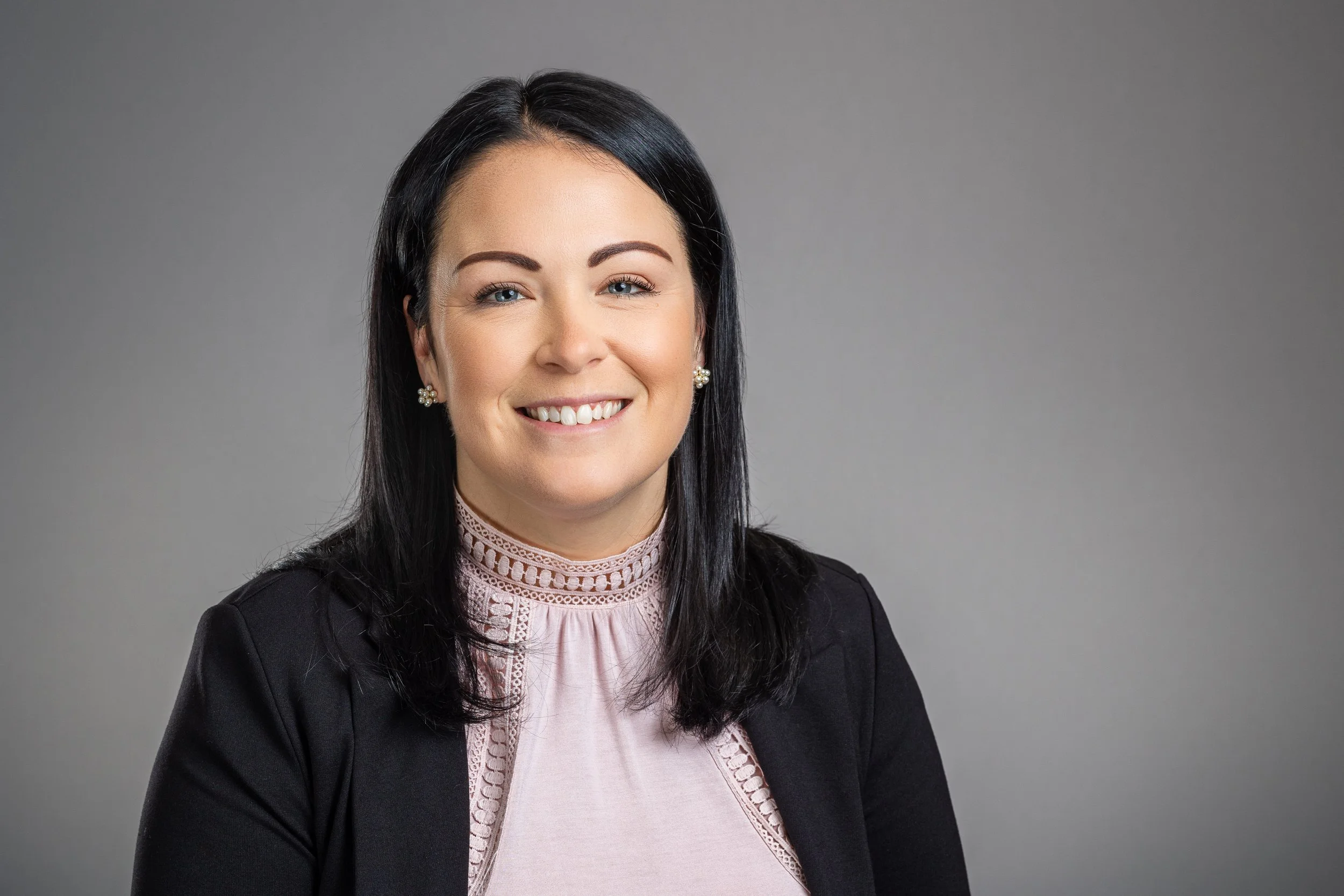 A woman with black hair, wearing a black blazer over a light pink blouse with lace details, smiles against a gray background.
