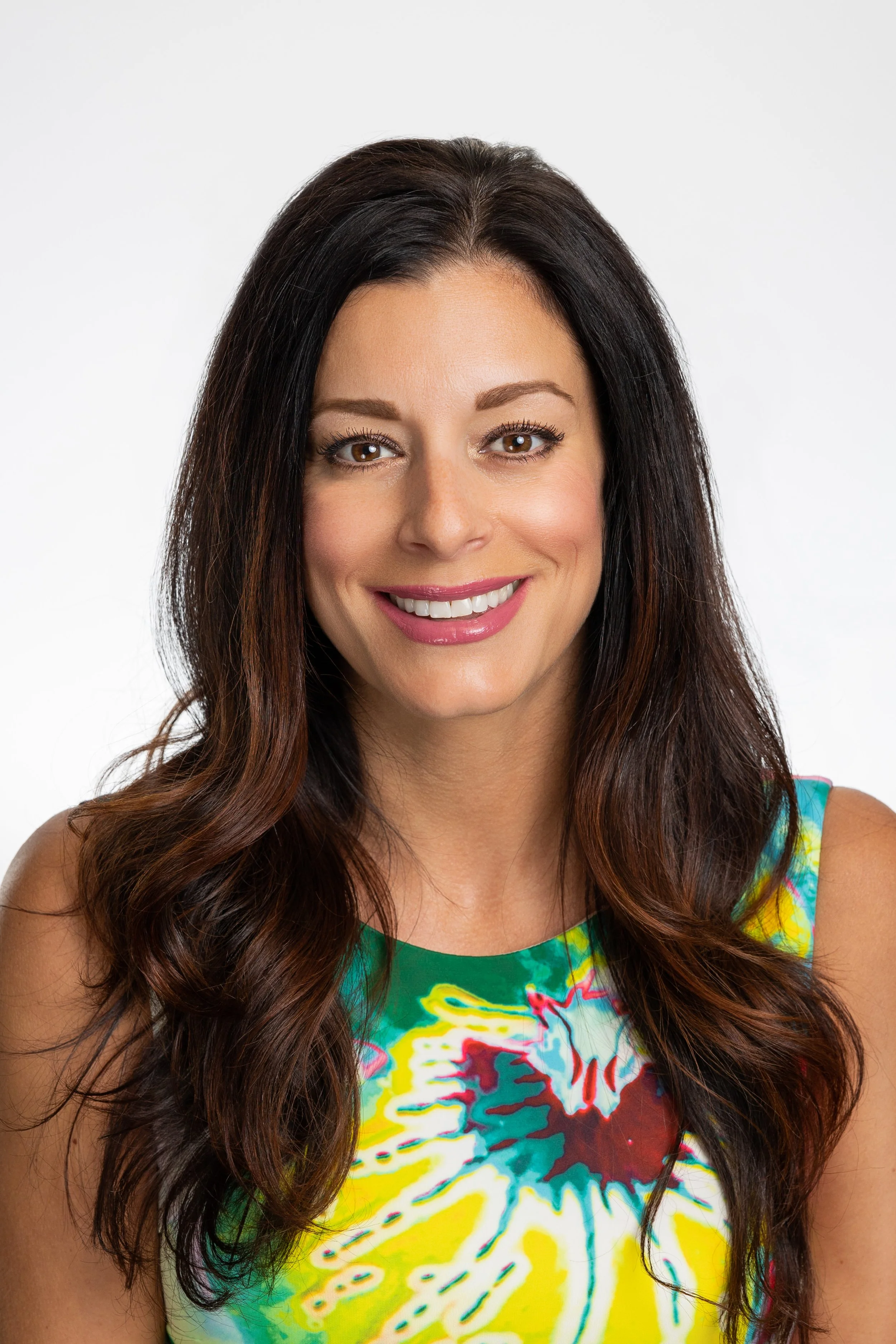 A smiling woman with long, wavy brown hair wearing a colorful tie-dye sleeveless top.