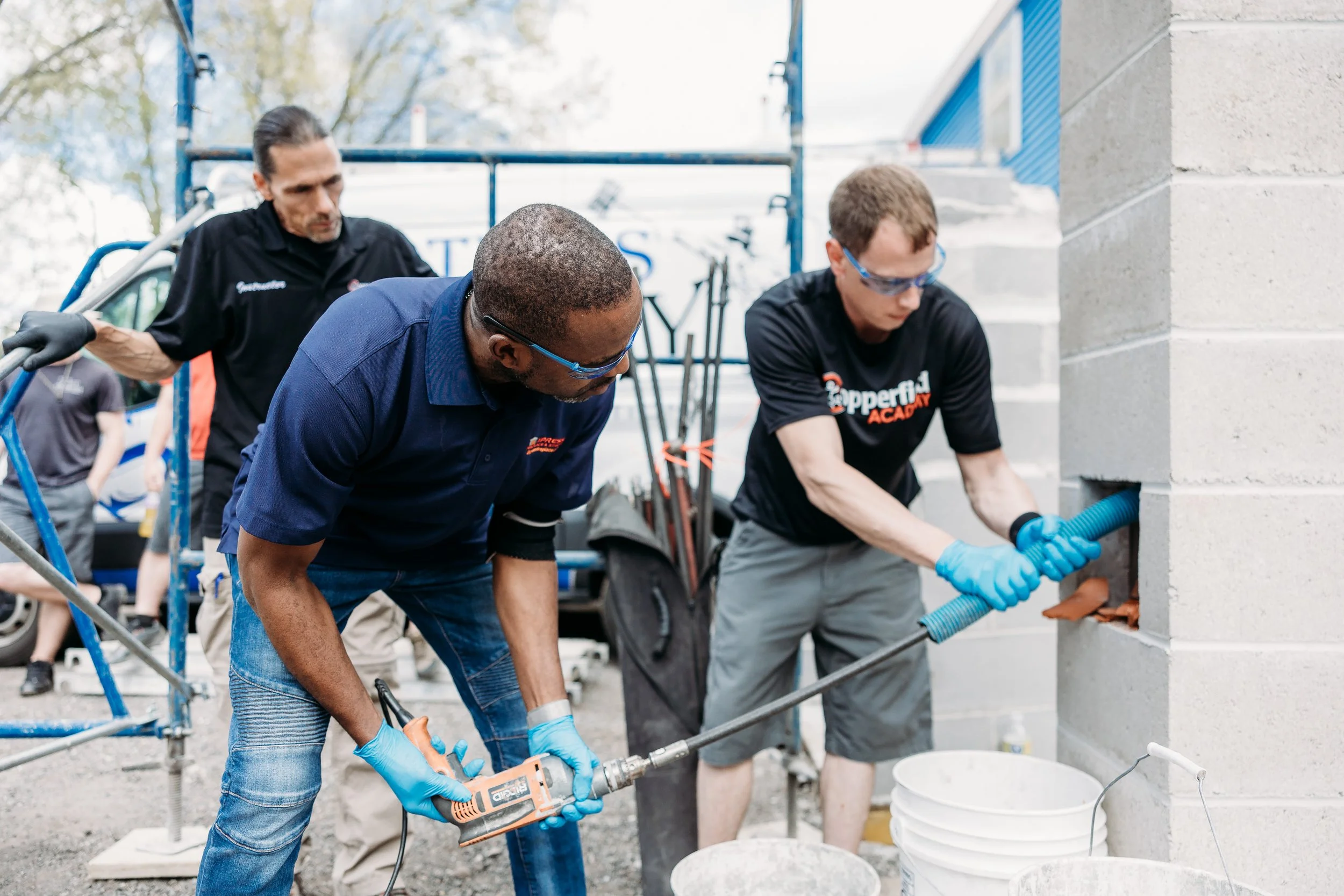 Three men working on a construction project outdoors, with one drilling into a concrete wall, the other using a pipe wrench, and the third in the background, all wearing gloves.