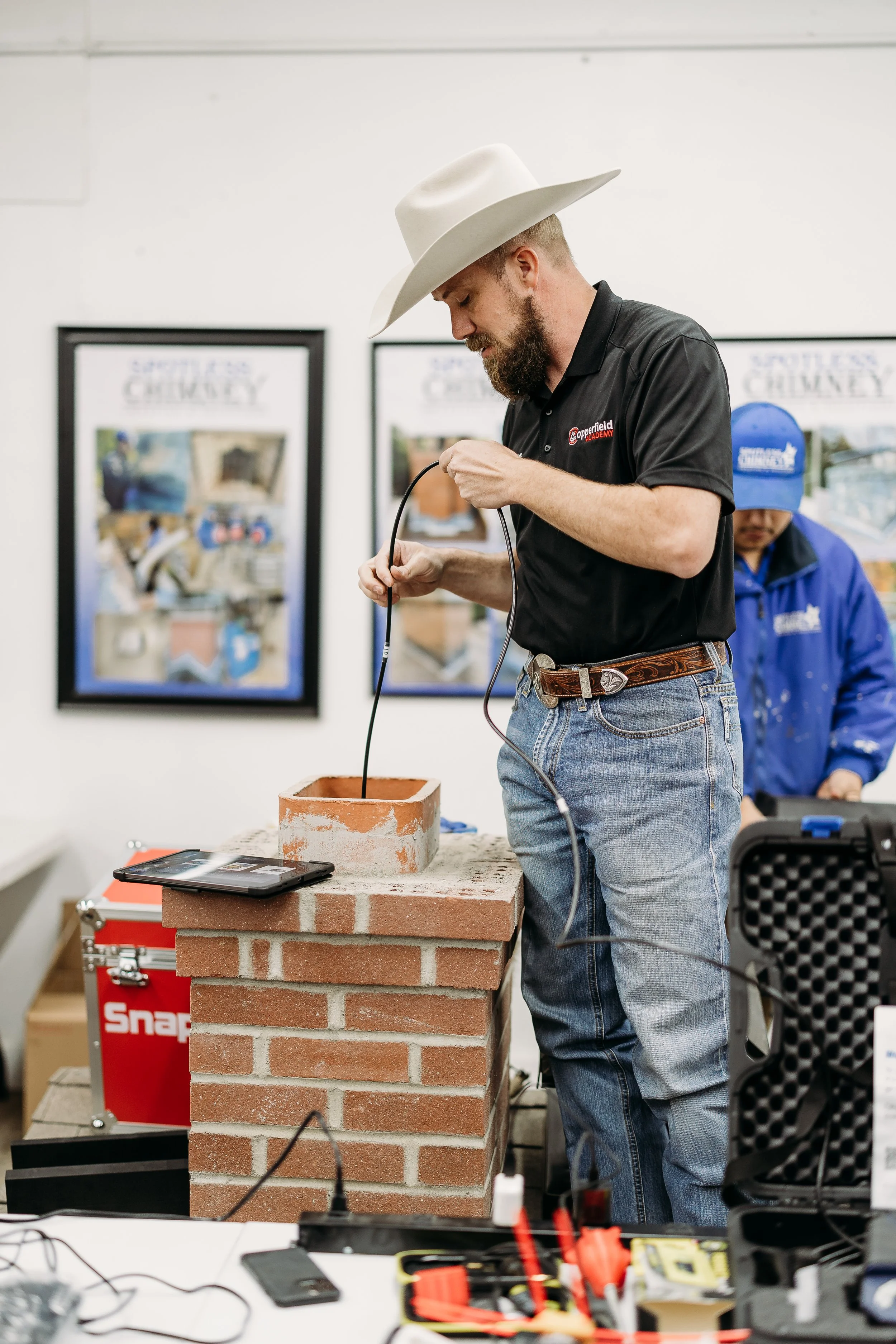 A man wearing a cowboy hat, black polo shirt, and jeans is conducting a science experiment involving a brick chimney and a glass tube. He appears focused while working on the setup, with another person in a blue jacket and cap in the background, in a