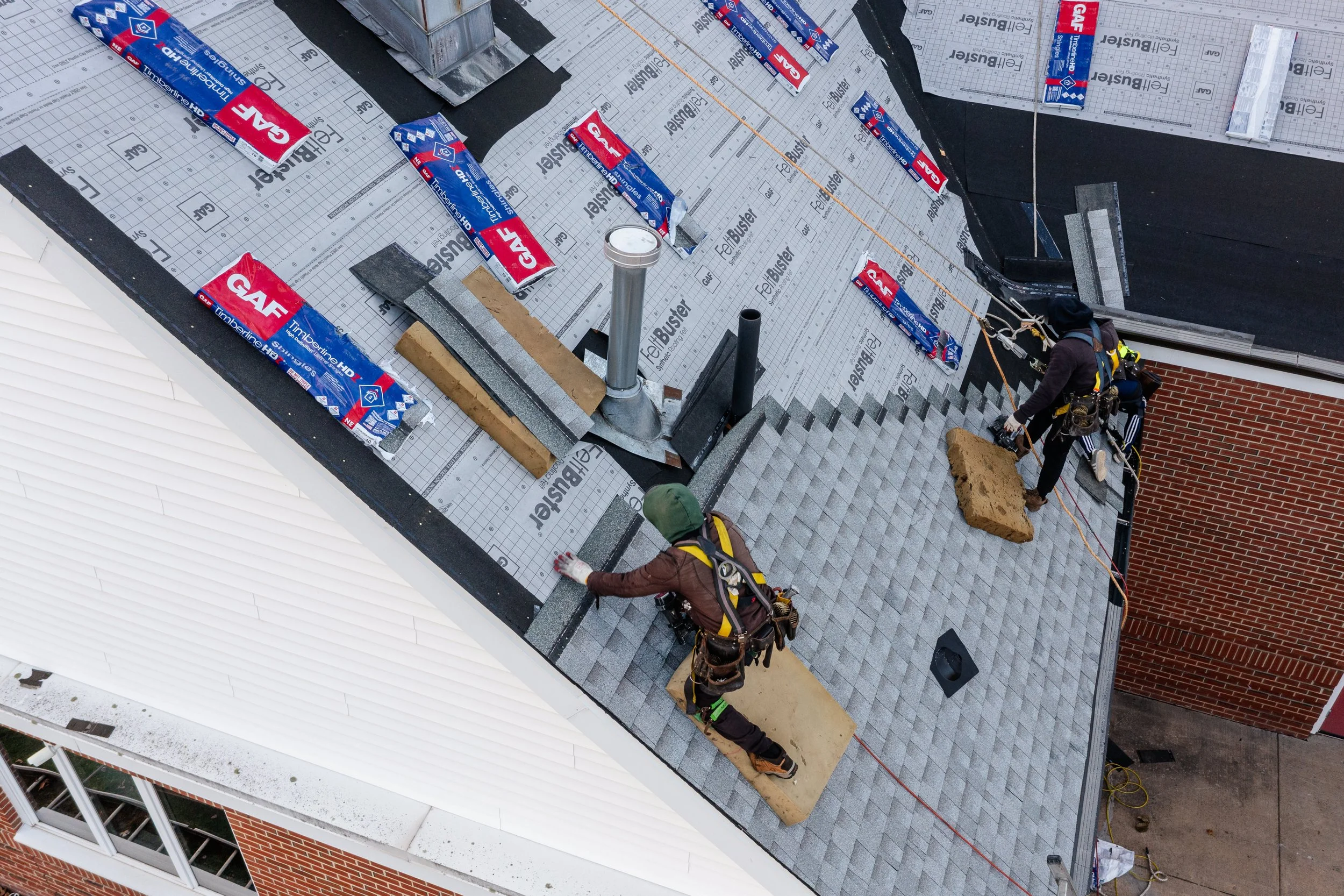 Two construction workers installing shingles on a roof, with one laying shingles and the other adjusting or securing them.