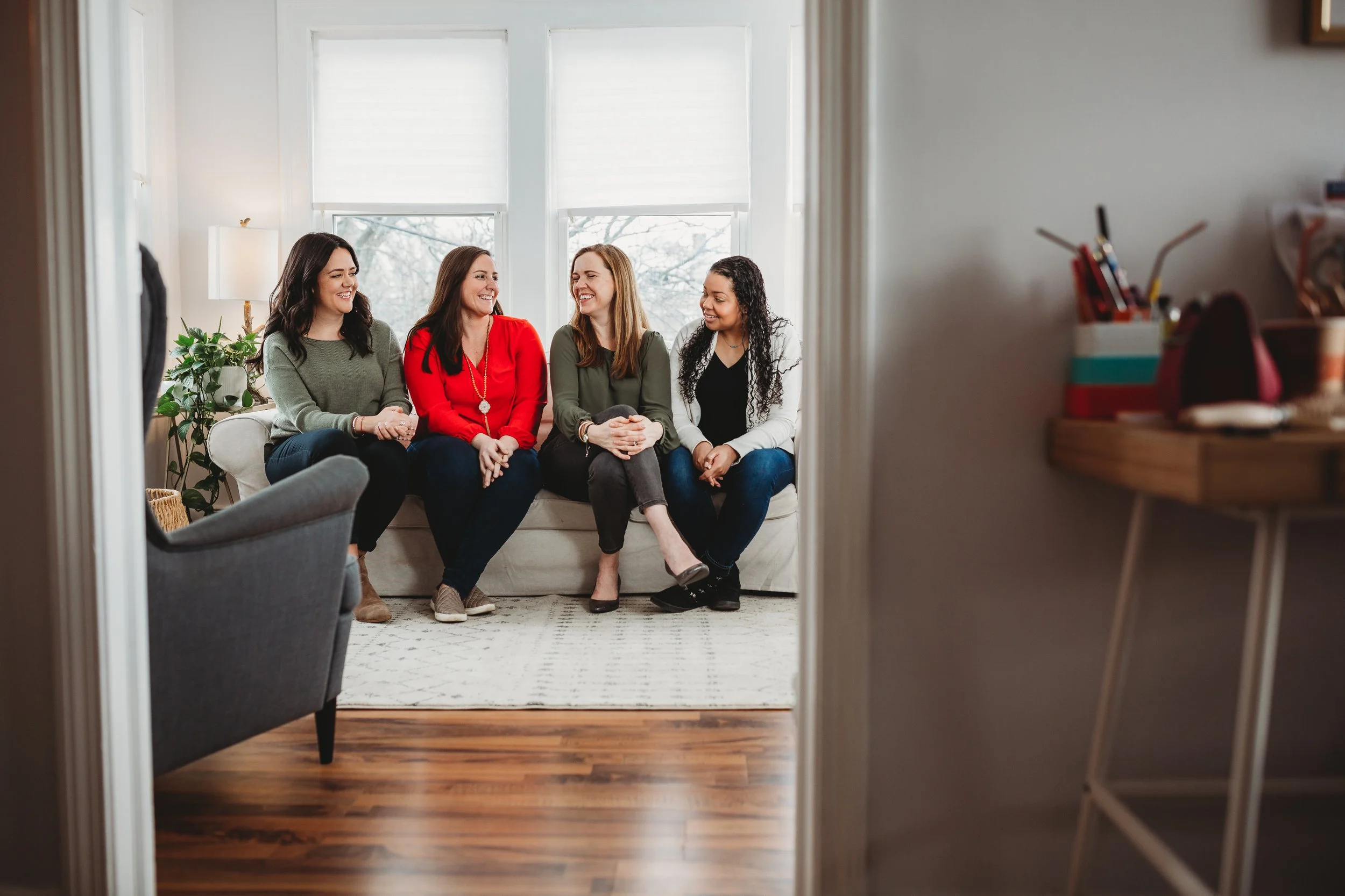 Four women sitting on a white couch, smiling and talking in a well-lit living room with large windows, a potted plant, and a patterned rug.