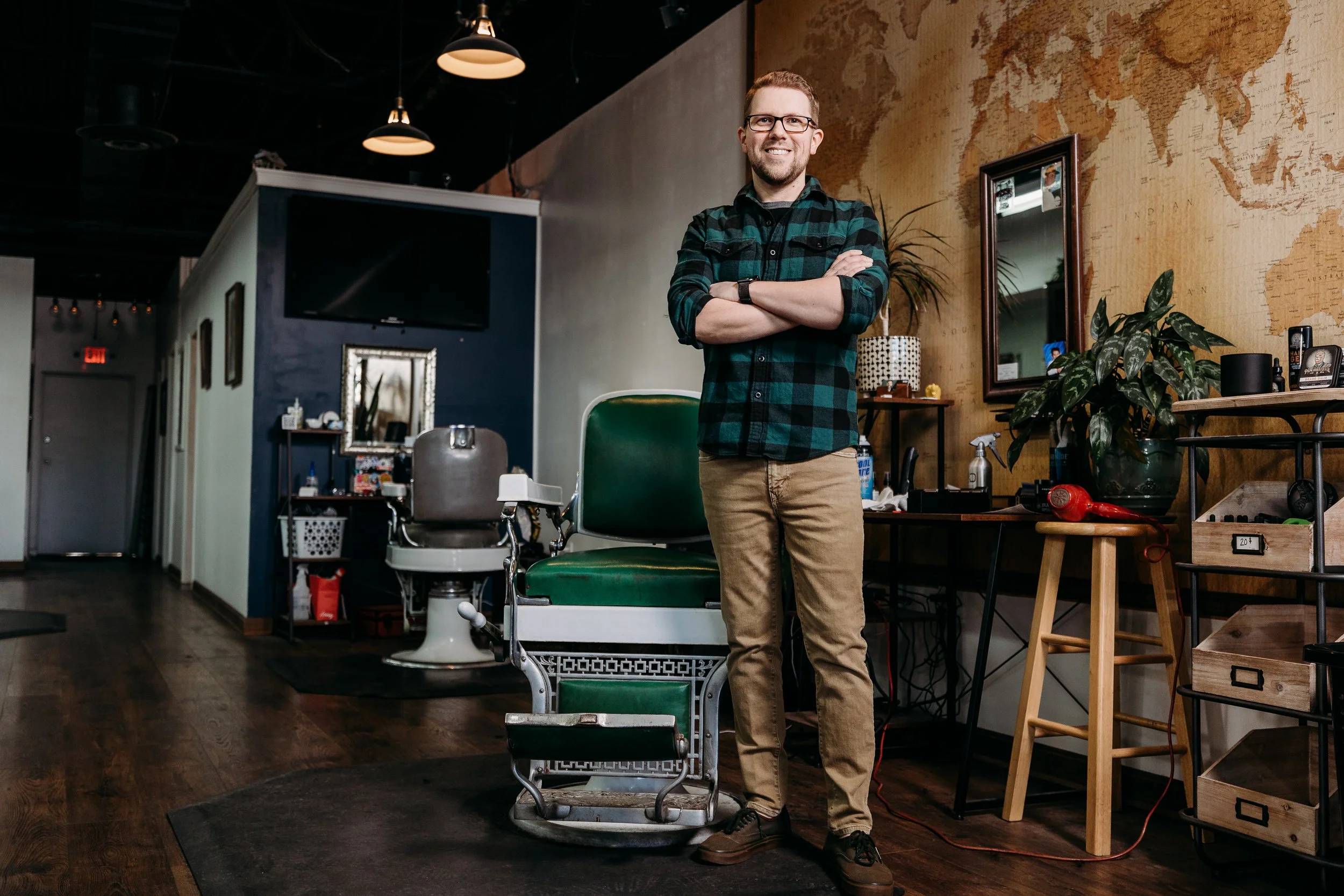 A man standing in a barbershop with arms crossed, wearing glasses, a green and black plaid shirt, beige pants, and brown shoes, smiling at the camera. The barbershop has a vintage barber chair, a large world map on the wall, a mirror, plants, and var