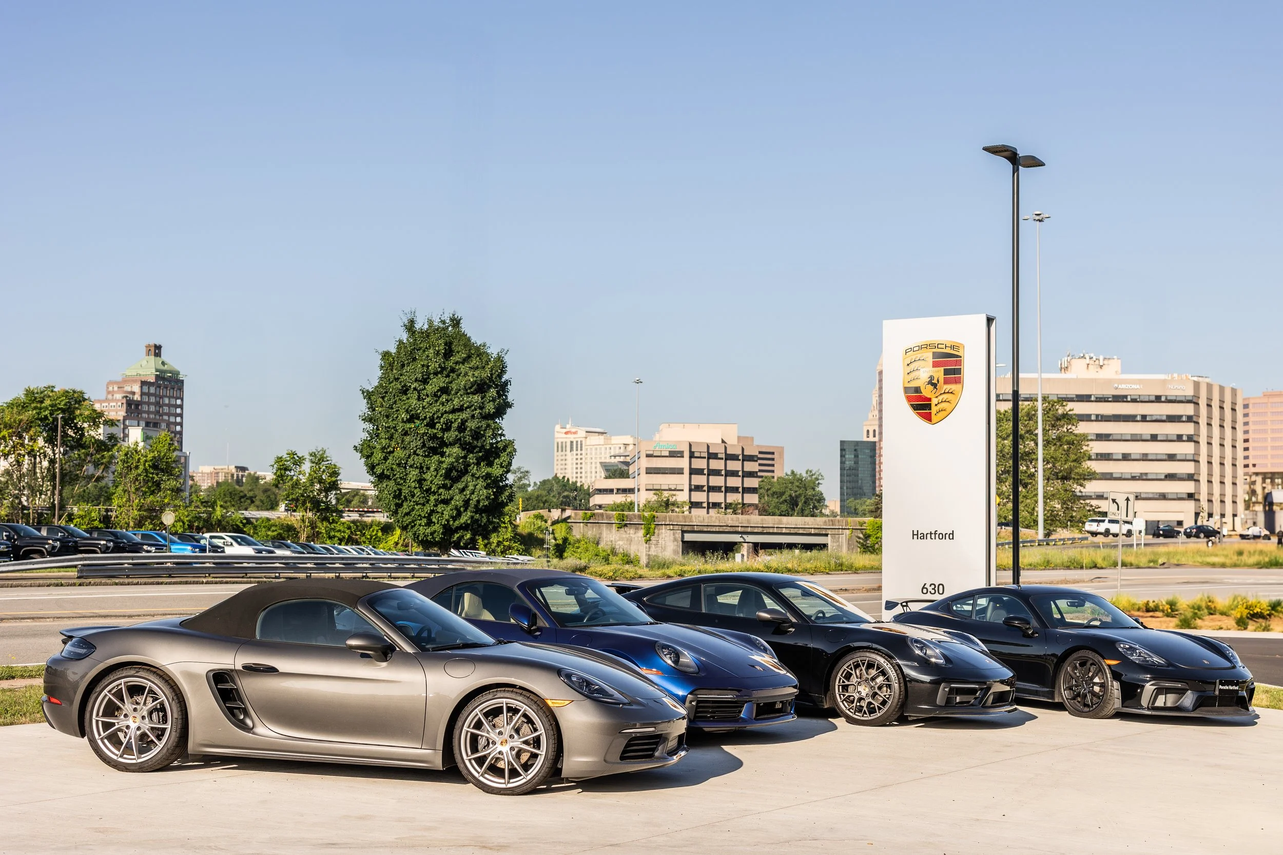 Four Porsche cars parked in front of a Porsche dealership sign that reads 'Hartford, 630' against an urban skyline with buildings, trees, and a clear blue sky.