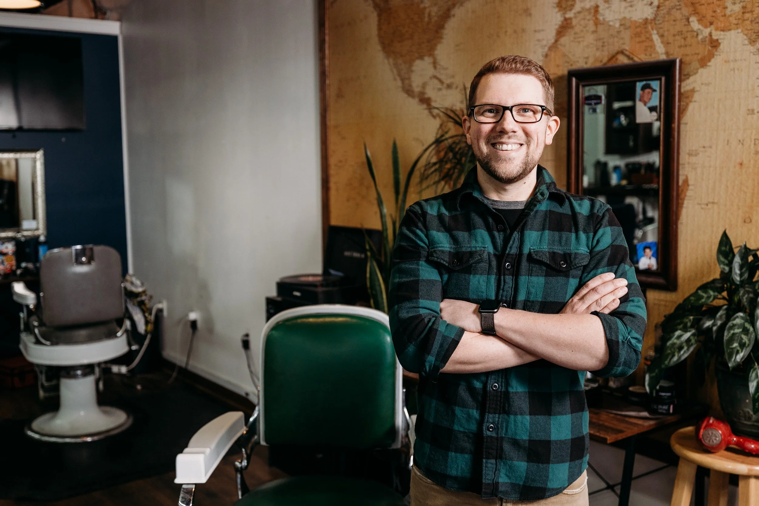 A smiling man with glasses and a beard standing with arms crossed in a barbershop or salon, with vintage chairs, a mirror, and plants in the background.