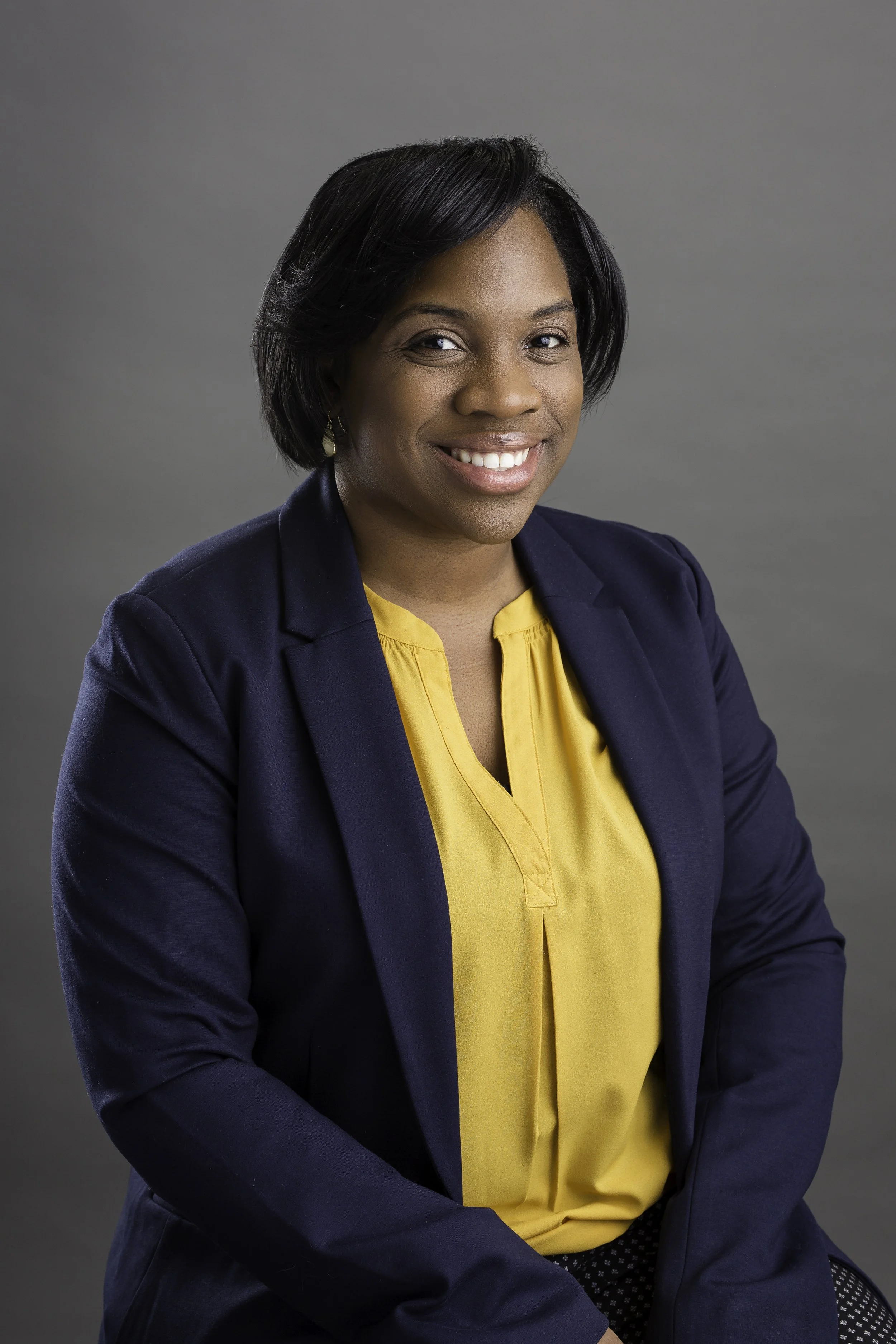 A professional woman with short black hair and earrings, wearing a navy blue blazer and a yellow blouse, smiling against a plain gray background.