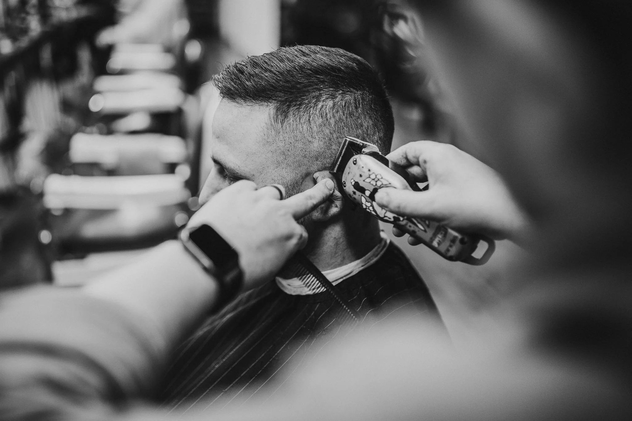A person getting a haircut, with a barber using electric clippers to trim the side of their head, while another person points at the haircut.