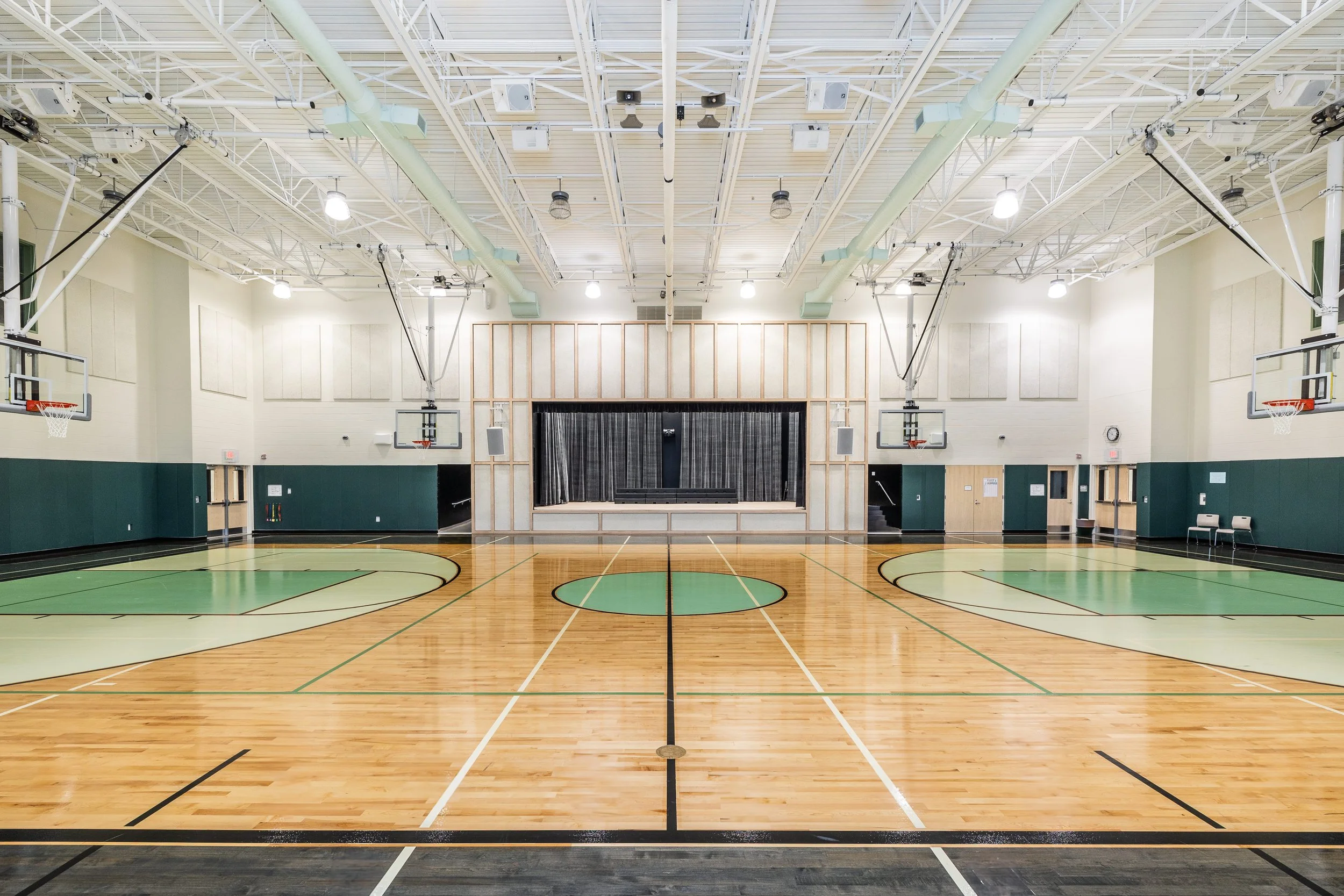 Empty indoor basketball gymnasium with wooden floor, basketball hoops, stage with curtains, and chairs, illuminated overhead.