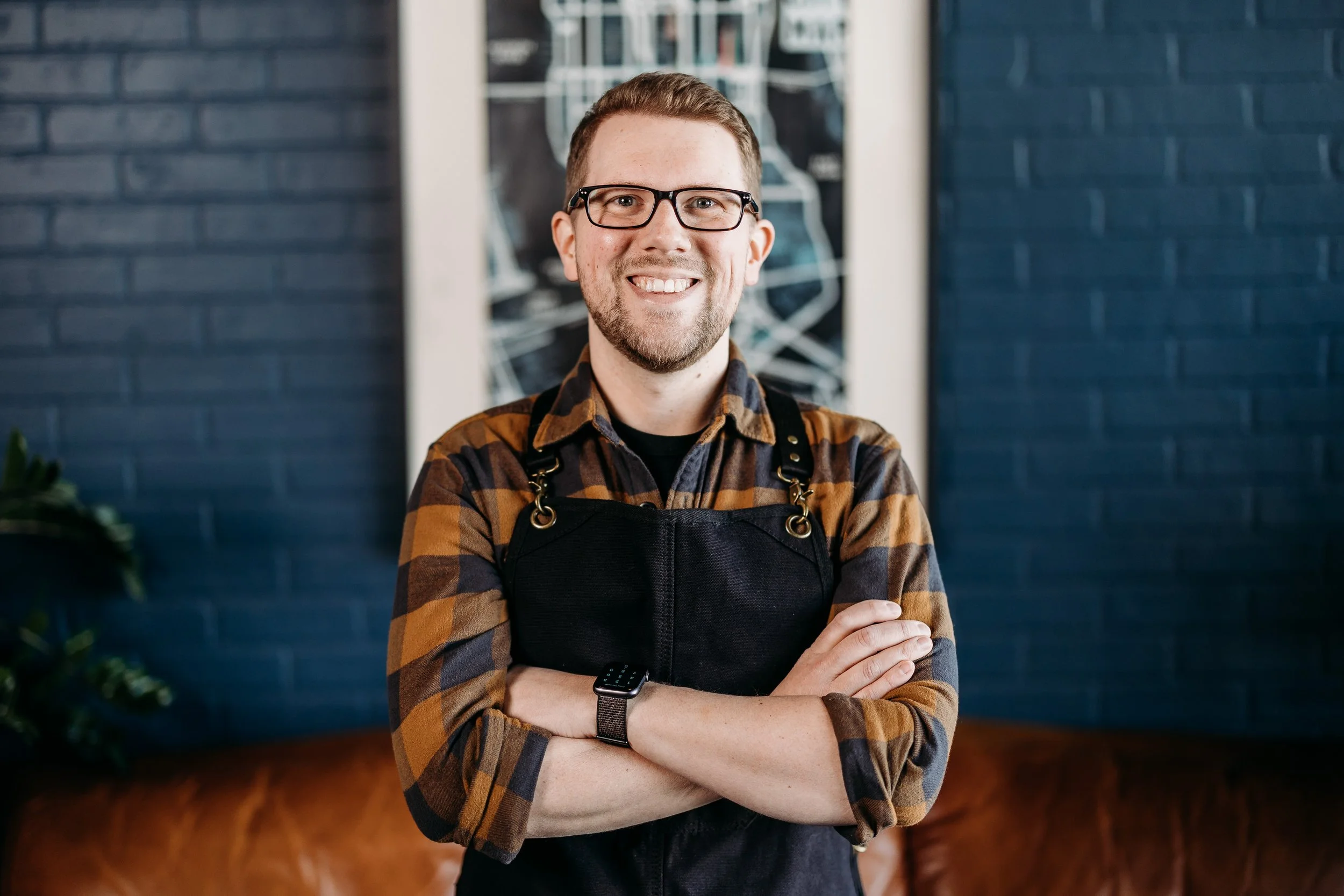 A smiling man wearing glasses, a black apron, and a brown checkered shirt inside a cafe or restaurant.