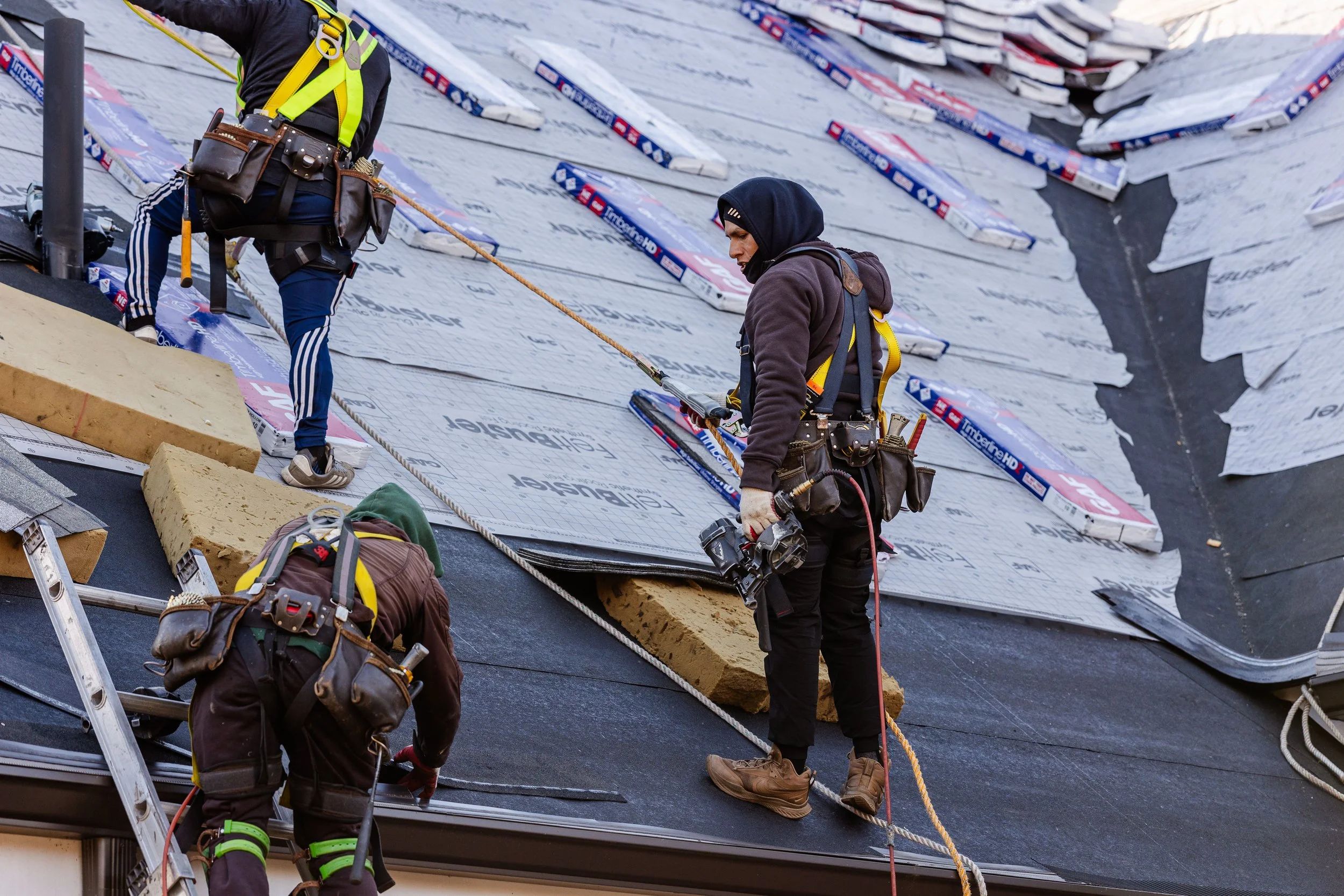 Three construction workers installing insulation foam on a sloped roof, with safety harnesses and tools.