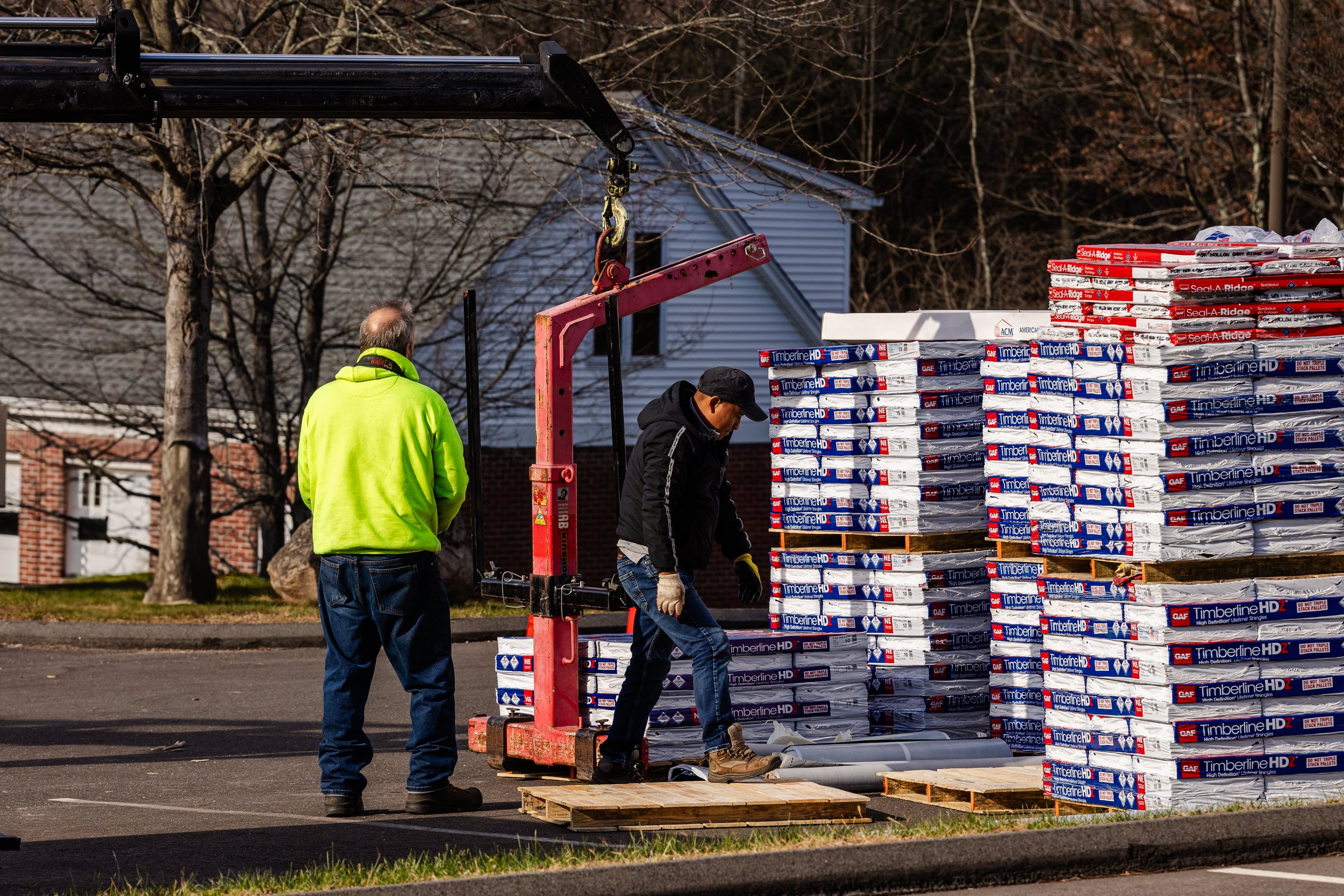 Two men working with stacks of rolls of roofing material labeled 'Timberline HD' and 'Seal-A-Ridge' outdoors, with one man in a black jacket and another in a bright yellow jacket, near a pallet and a construction crane.