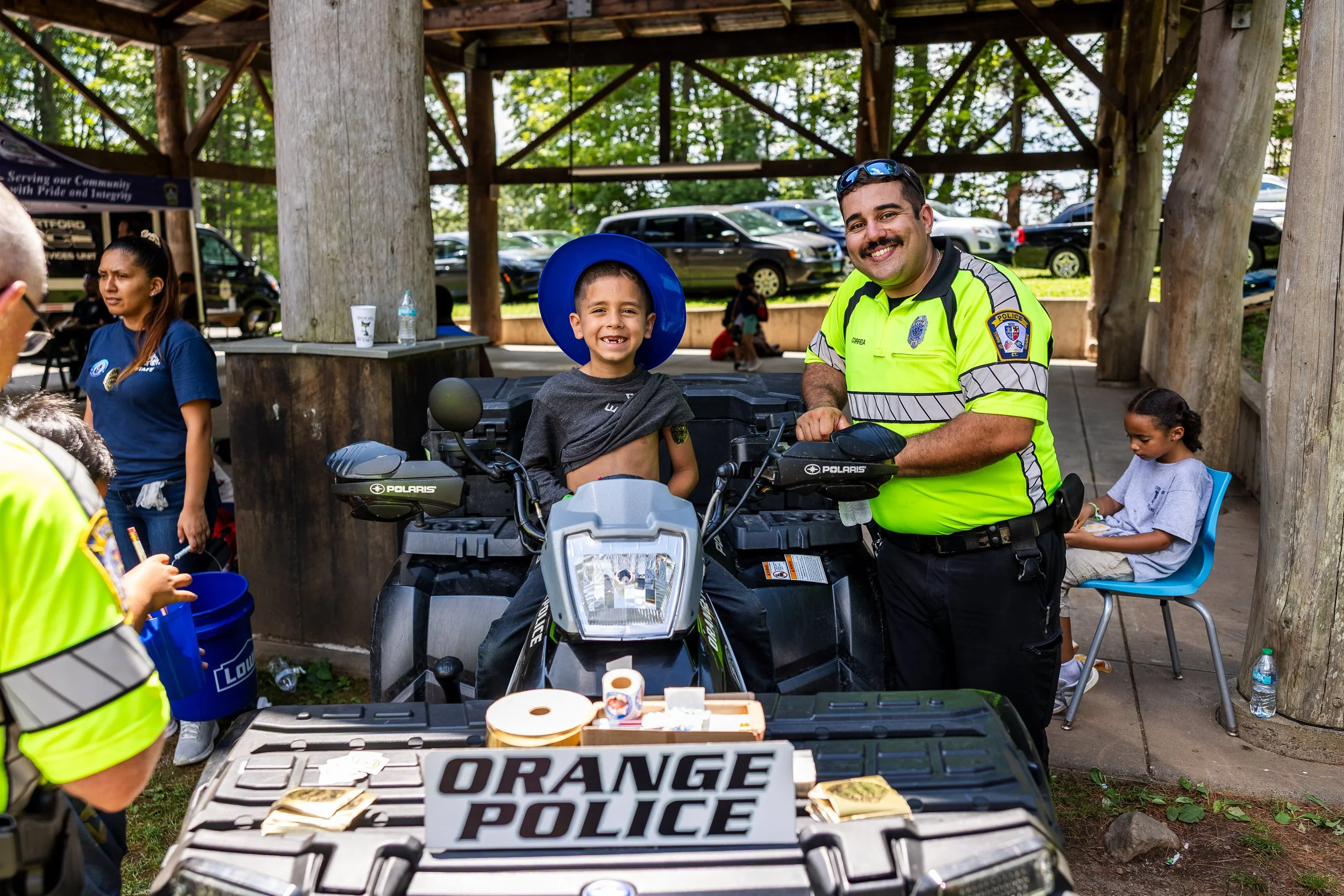 A young boy sitting on an ATV with a police officer at an outdoor community event. The boy is smiling and wearing a large blue helmet. The police officer, in a bright yellow uniform, is standing beside him, also smiling. There is a table in front of 