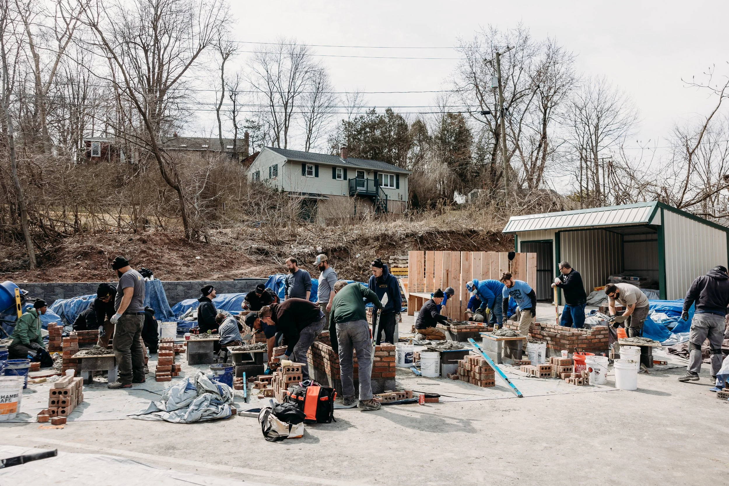 Group of people working together on a construction site building a brick wall, with tools and materials scattered around, outdoors in a residential area during daytime.