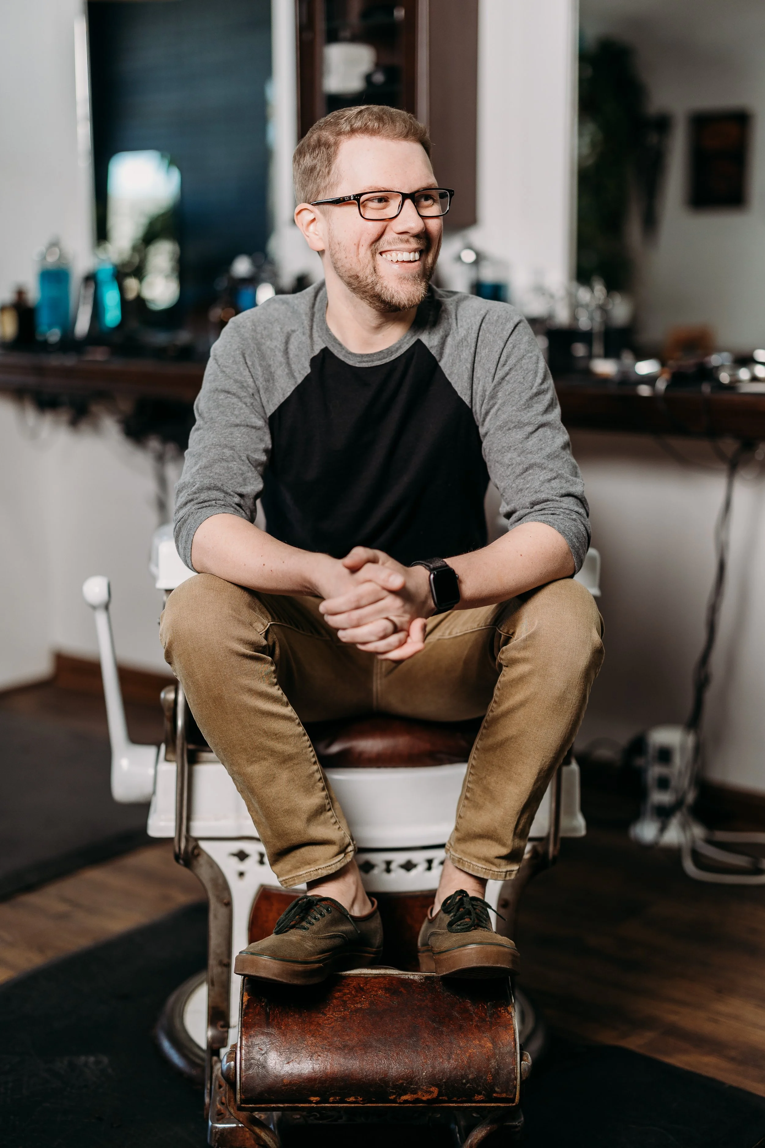 A man with glasses and a beard, smiling, seated on a vintage barber's chair in a barbershop, wearing a gray and black long-sleeved shirt, beige pants, and brown shoes.