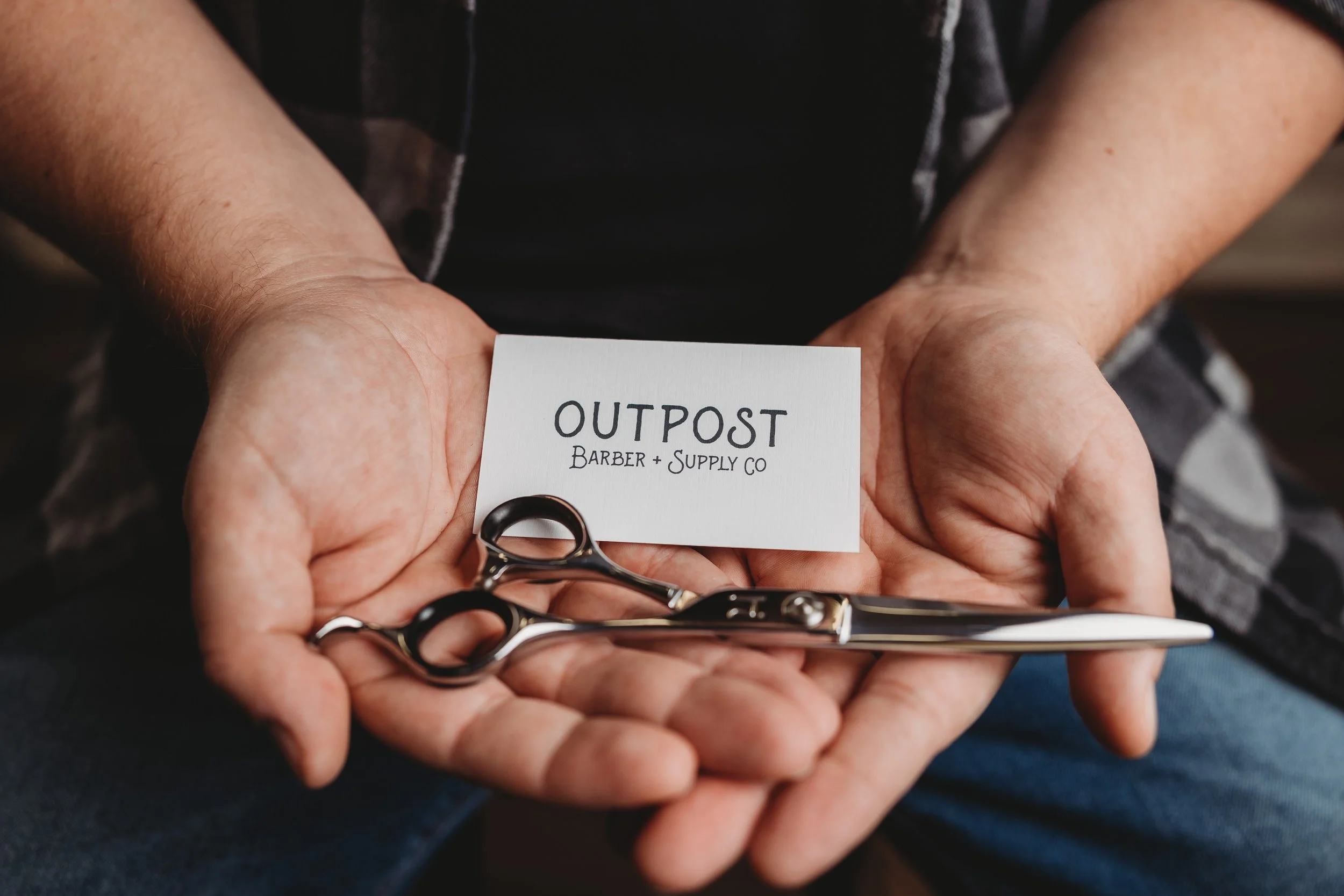 Person holding a pair of scissors, a business card that reads Outpost Barber + Supply Co, and some hairdressing tools.
