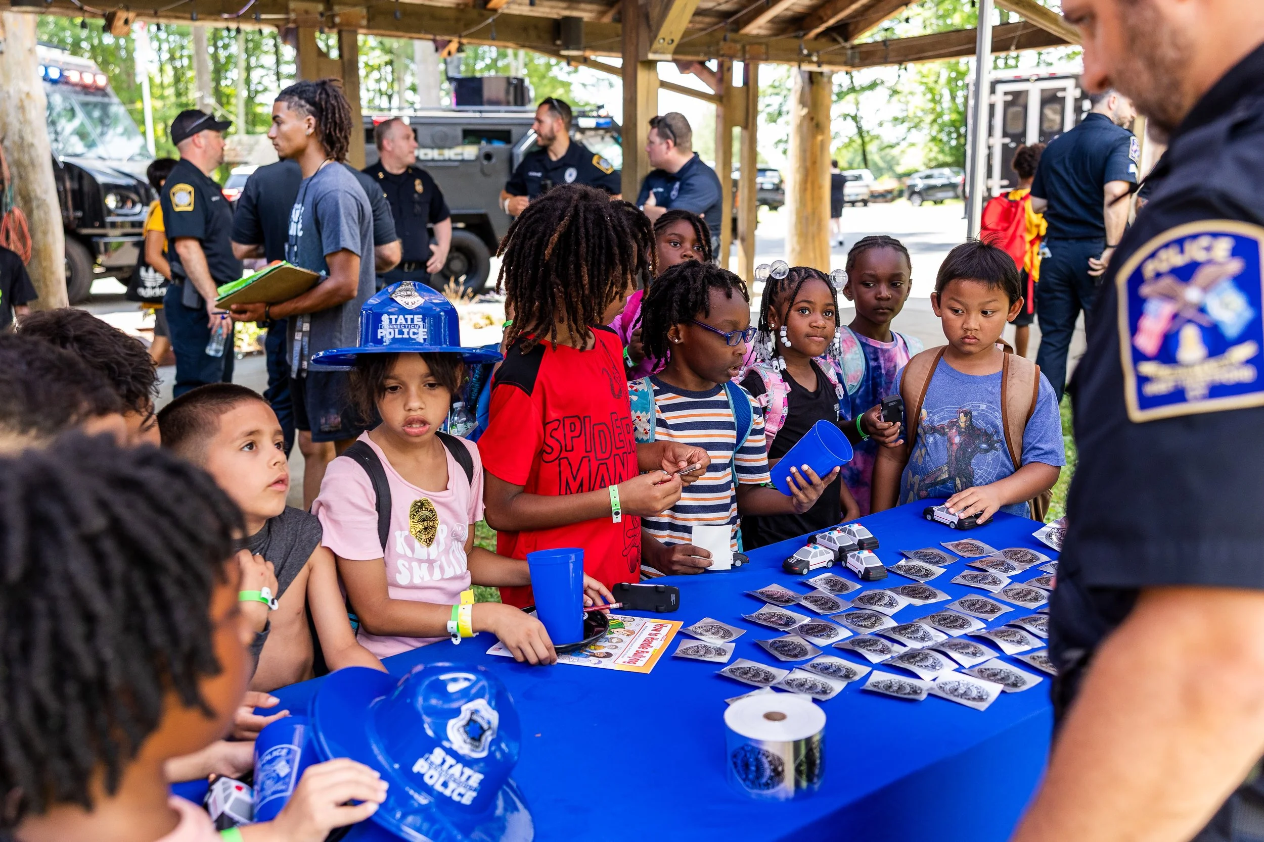 Children gathered around a blue table with police hats and badge stickers, talking to police officers at an outdoor community event.