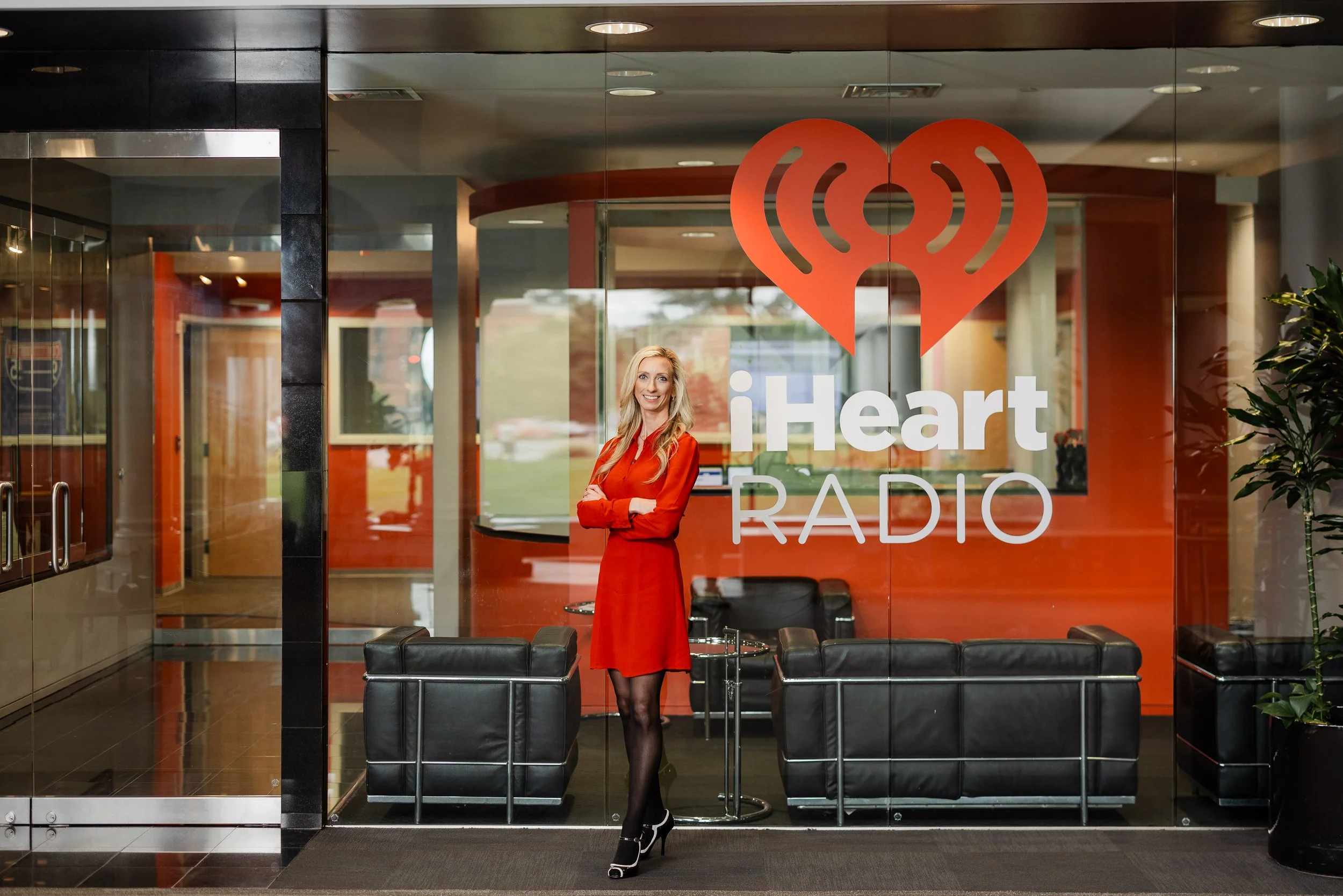 Woman in red dress standing in front of iHeartRadio studio window with logo, black chairs and glass entrance door