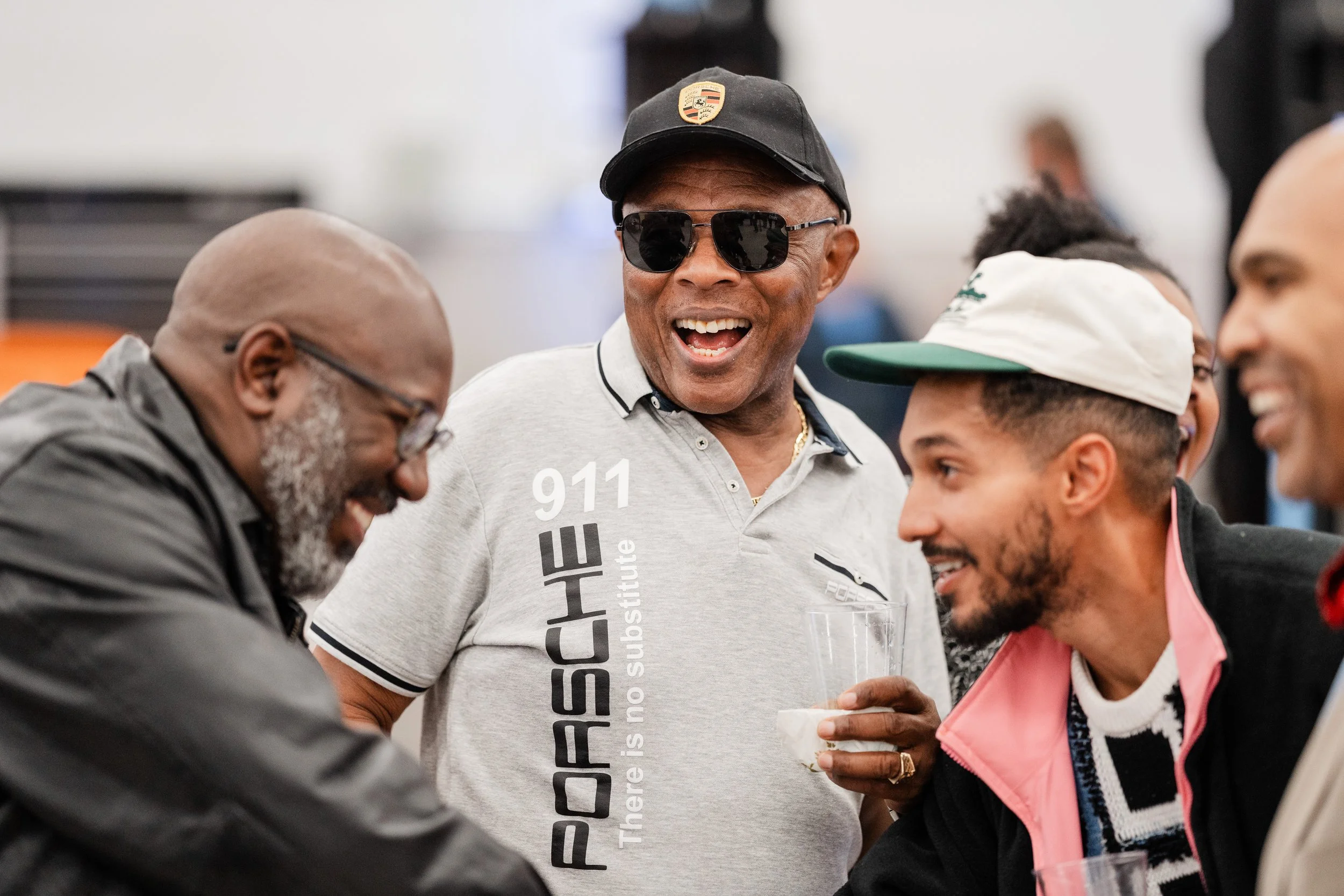 Group of four men smiling and talking at a social gathering, one wearing sunglasses and a Porsche shirt, holding a drink.