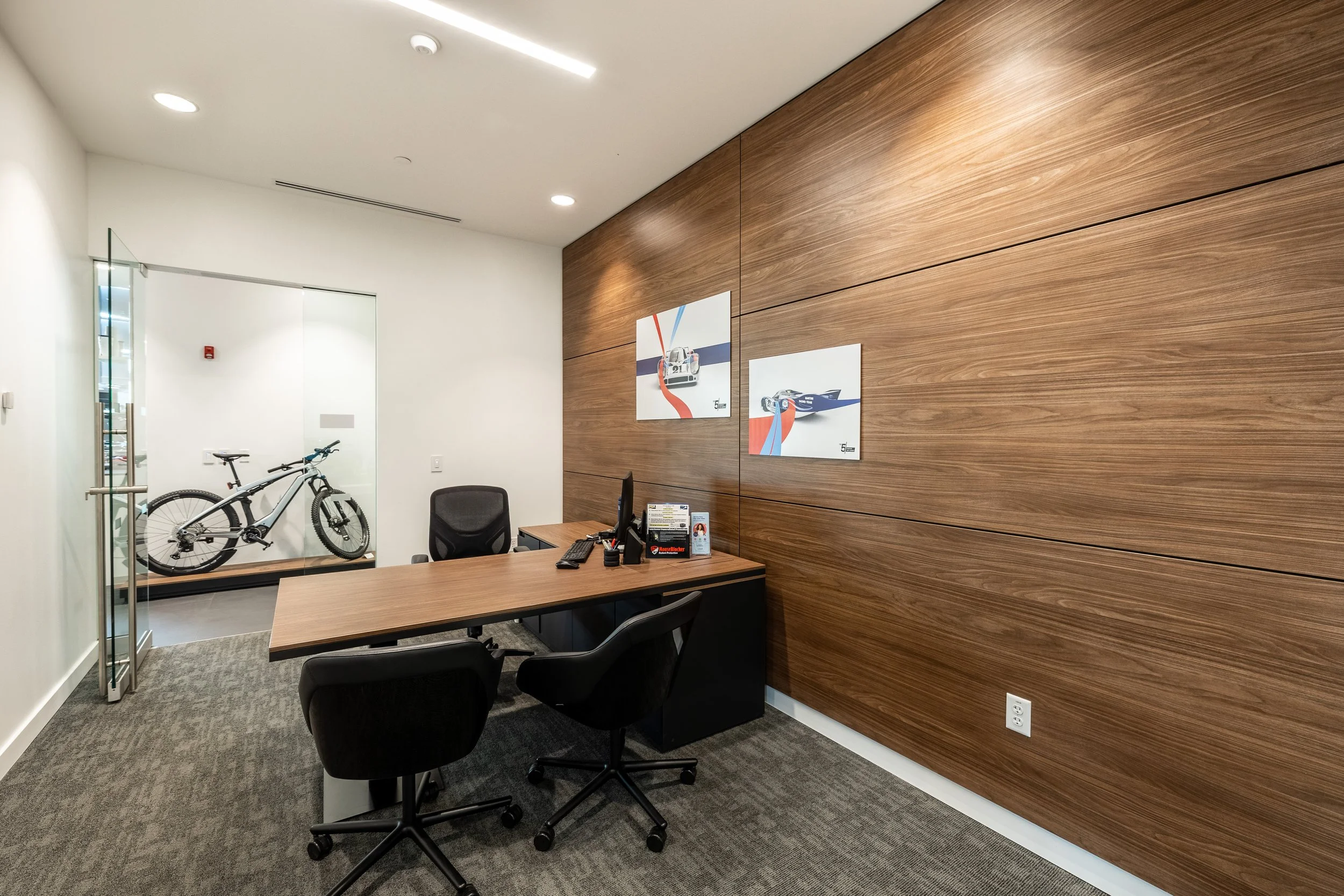 Modern office reception area with a wooden wall, artwork, a desk with a computer, and a glass wall showcasing a bicycle.