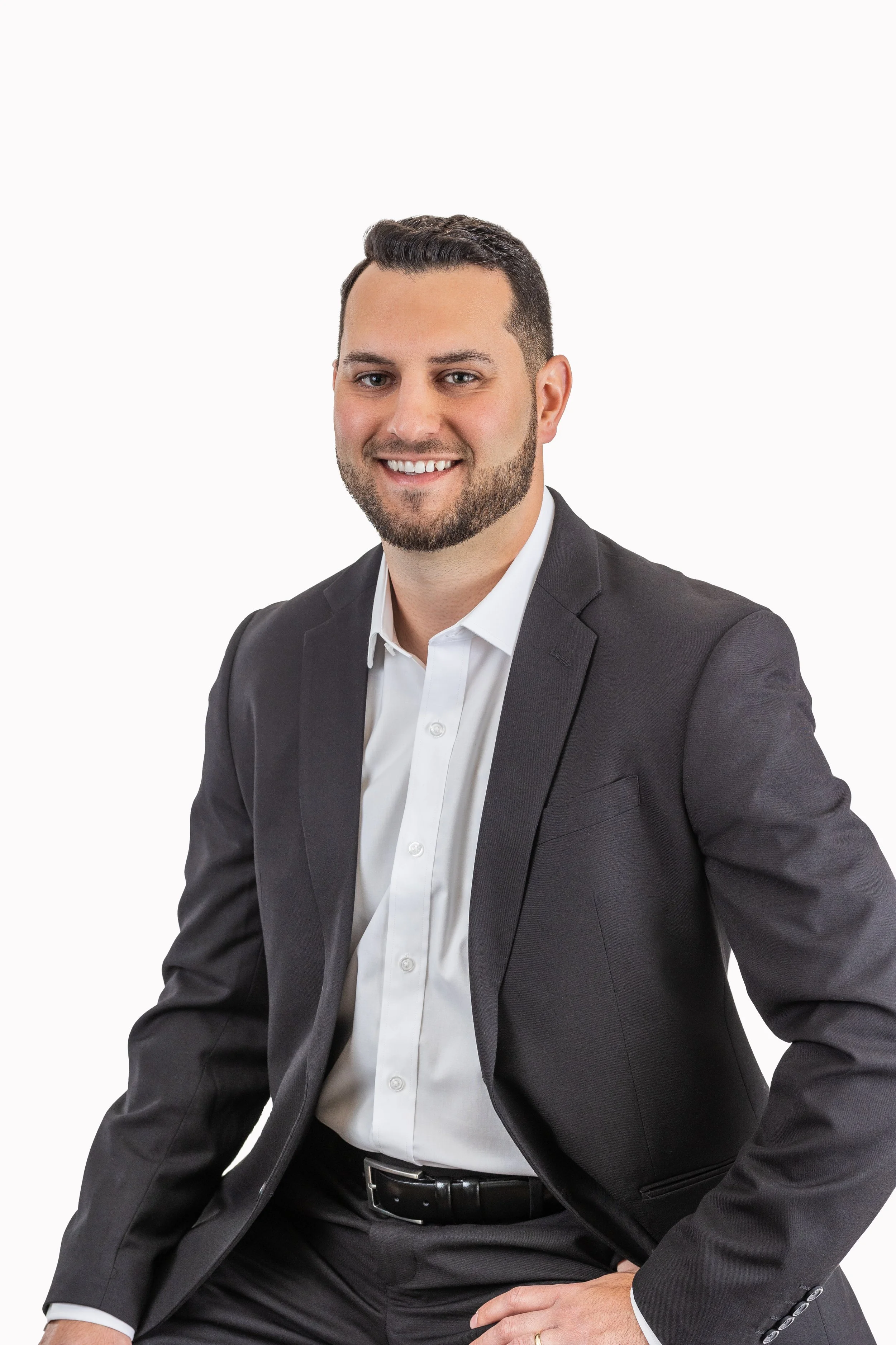 Portrait of a man with dark hair, a beard, smiling, dressed in a black suit and white shirt, sitting against a plain white background.