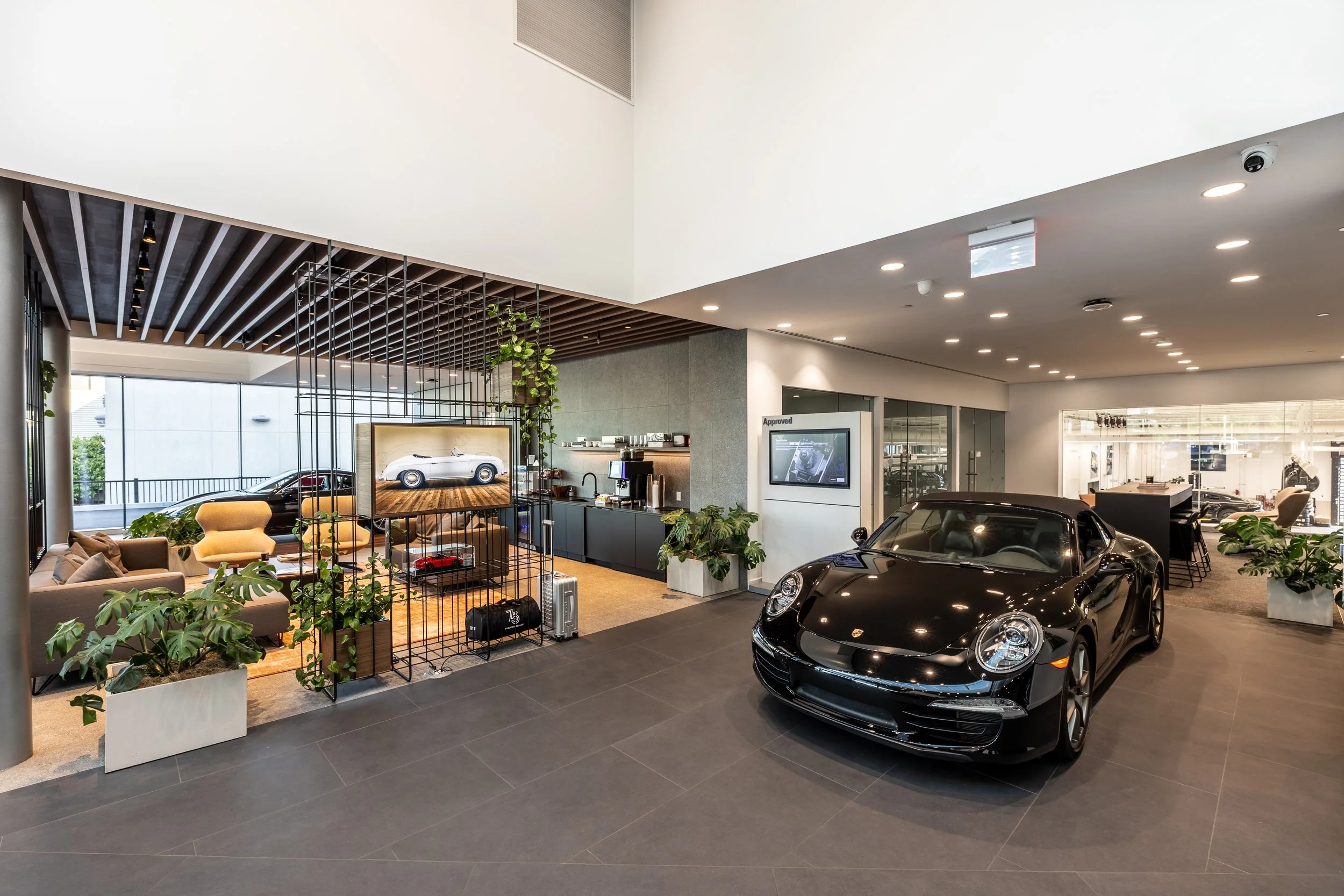 Modern car dealership showroom with black sports car, seating area, large window, potted plants, and a coffee station.